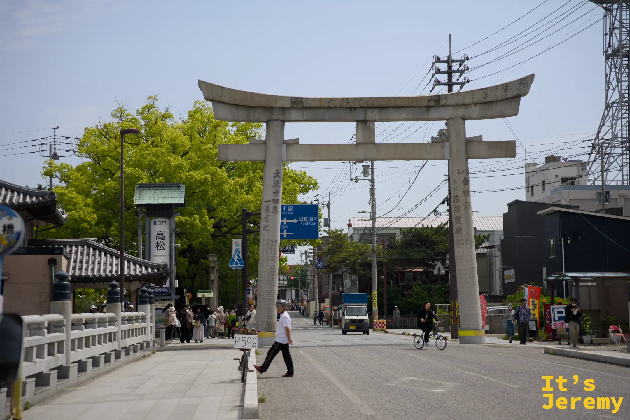 琴平大鳥居矗立在表參道入口路口，石造鳥居後方可見琴電高松方向指示牌與綠色大樹