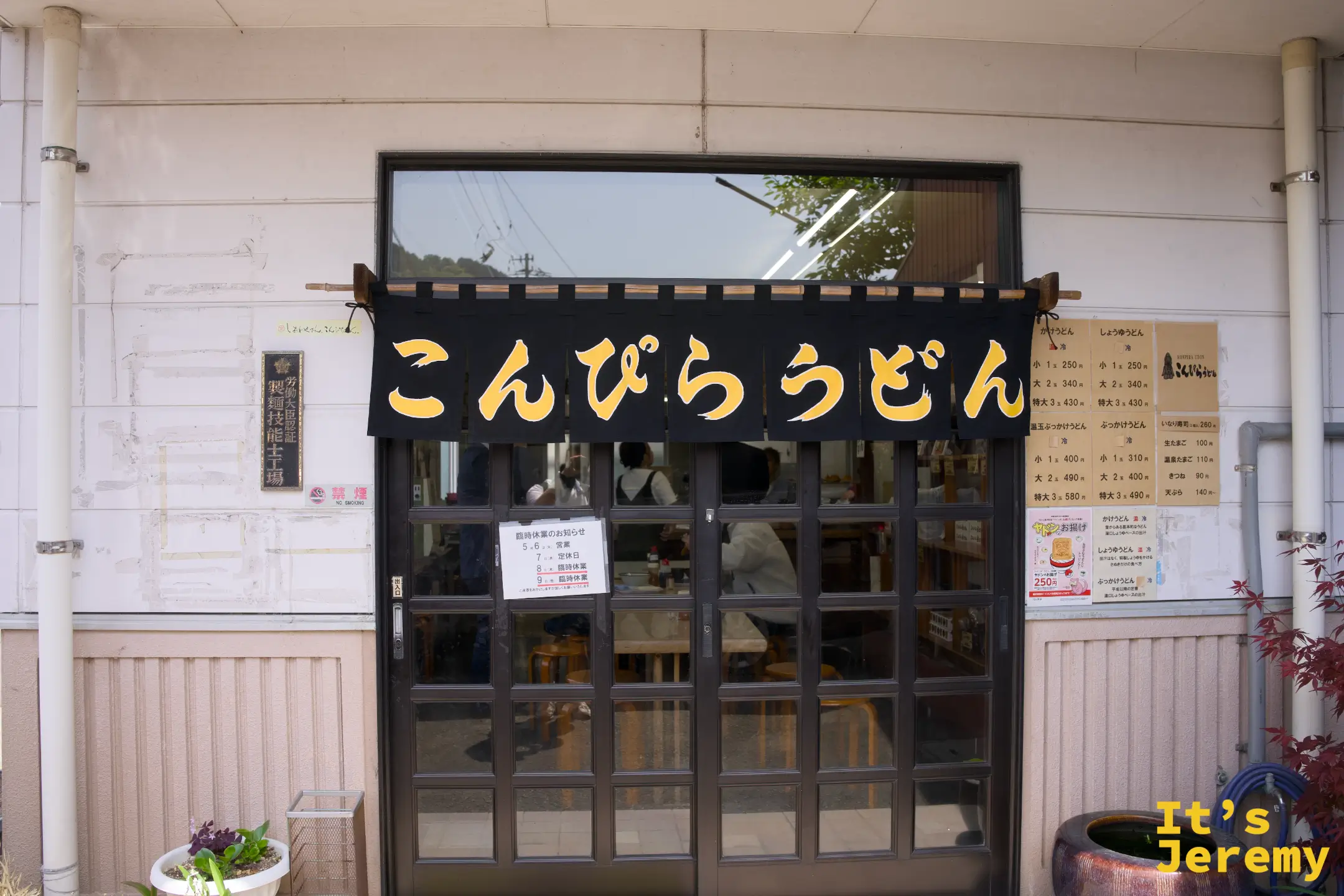 こんぴらうどん 工場店正面，黑色暖簾上寫著金黃大字「こんぴらうどん」，旁邊牆上掛著製麵所的認定牌
