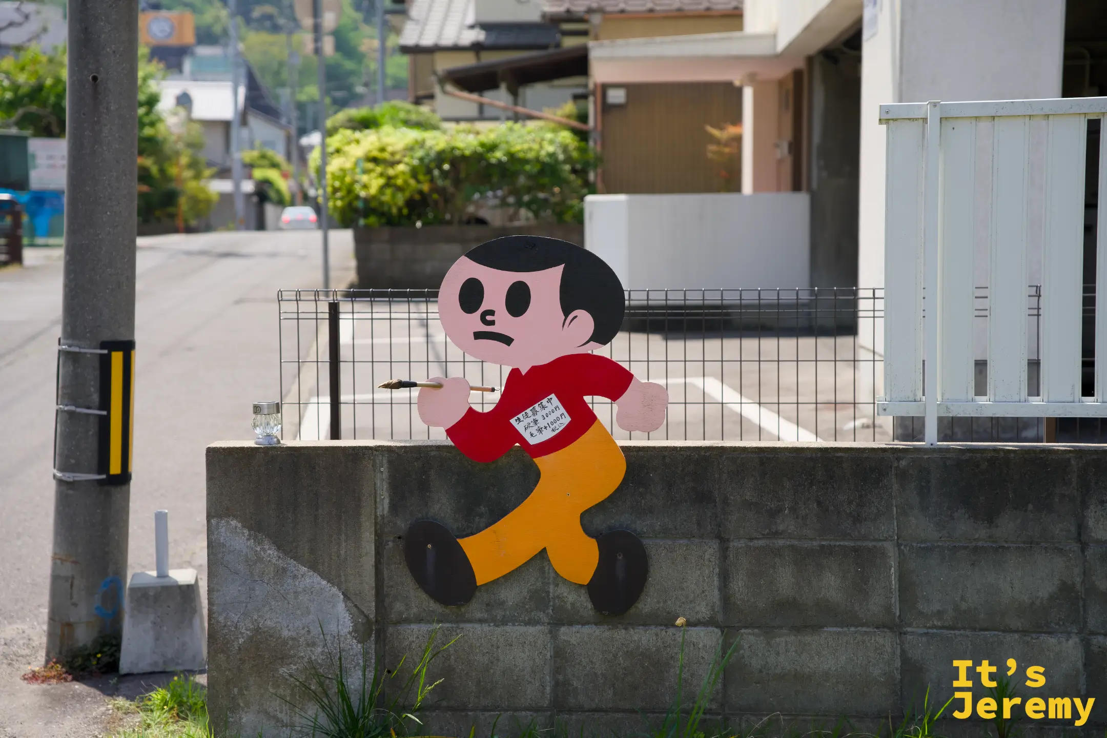 橘紅色飛び出し坊や立體剪影人形固定在住宅區圍牆邊緣，背景是安靜的住宅小路