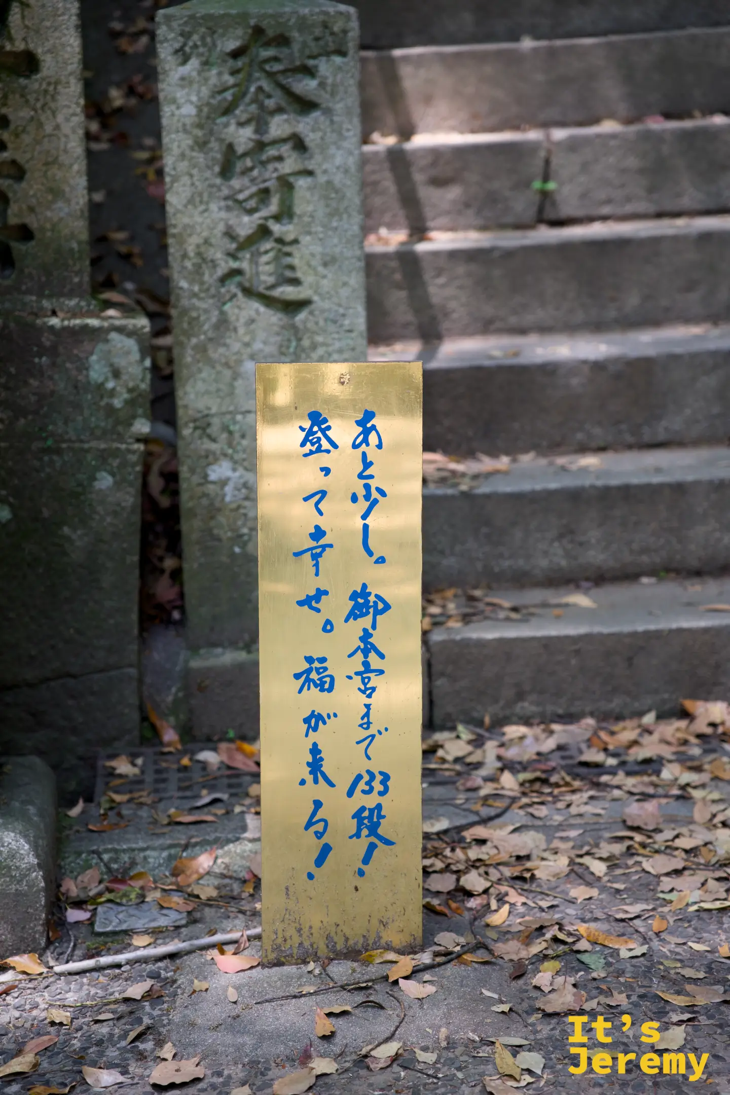 金色立牌上用藍色毛筆字寫著「あと少し。御本宮まで133段！登って幸せ。福が来る！」，旁邊是落葉覆蓋的石階
