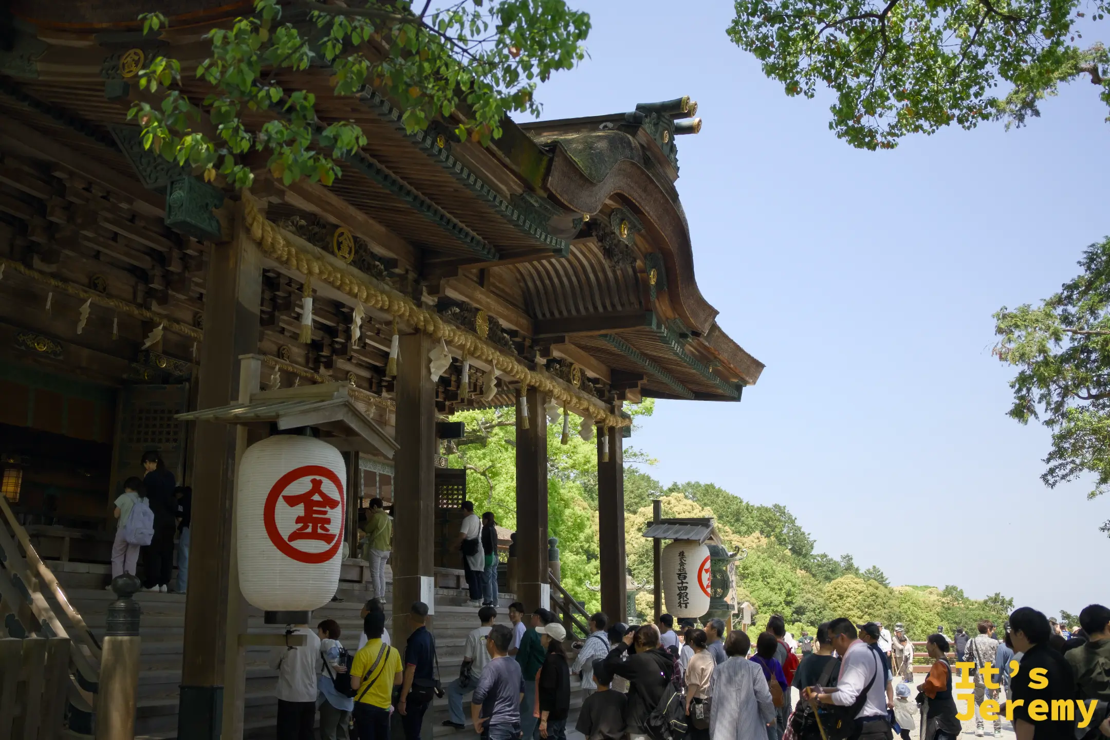 御本宮正面橫向景觀，參拜者在廣場和殿前聚集，金丸印燈籠懸掛在長廊廊柱上