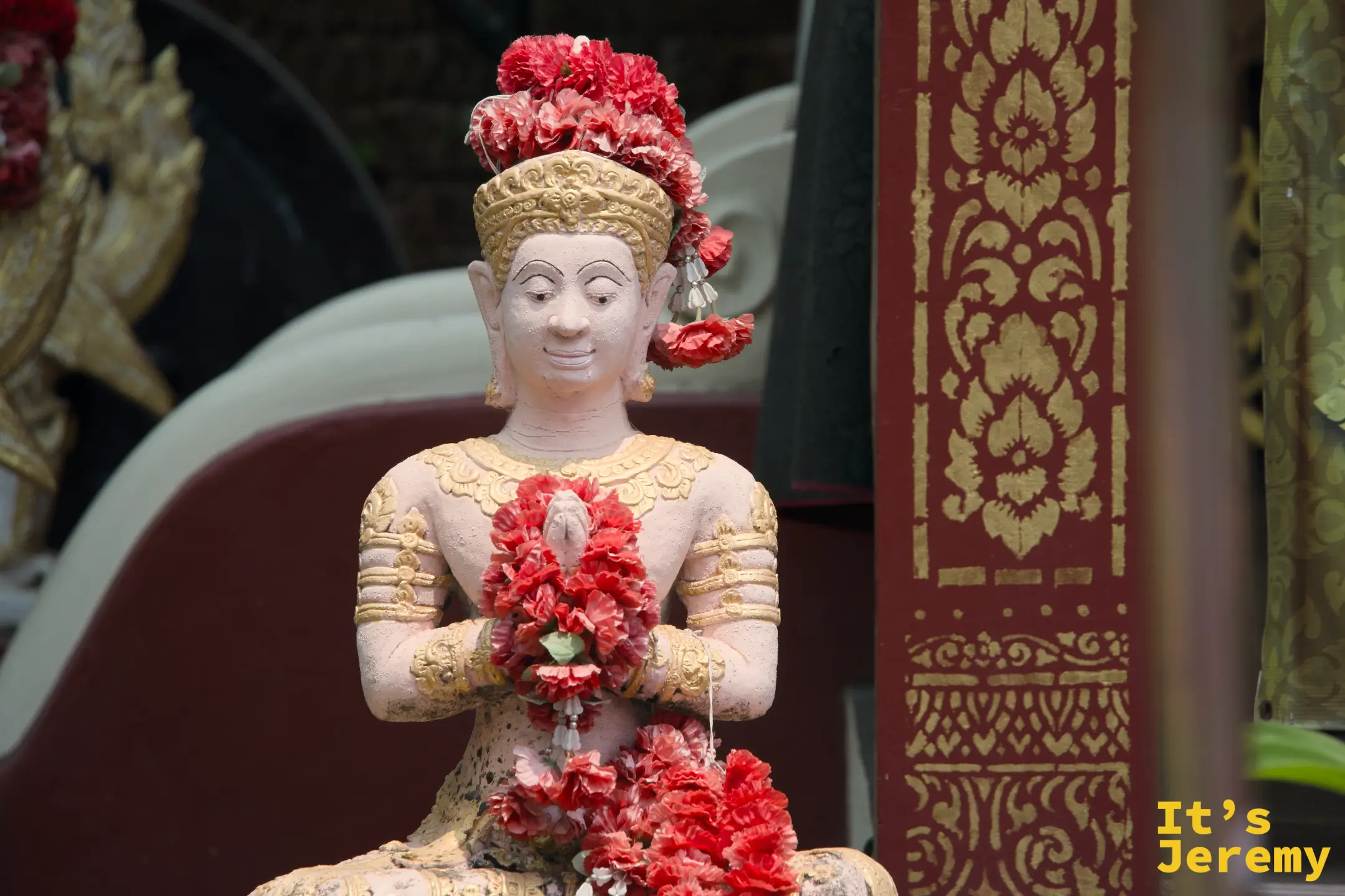 合掌祈願的男性天神像，頭戴紅色花冠，雙手捧著花環，神情專注，背景是金紅色的廟堂裝飾