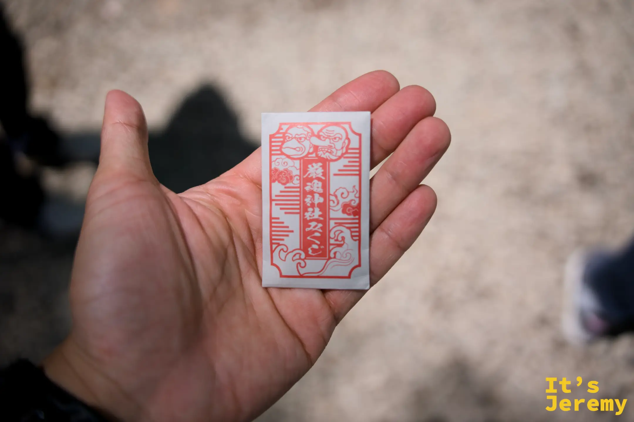 手心平攤，展示厳魂神社的護符，橘紅色設計印有神社名稱和兩位神明圖像