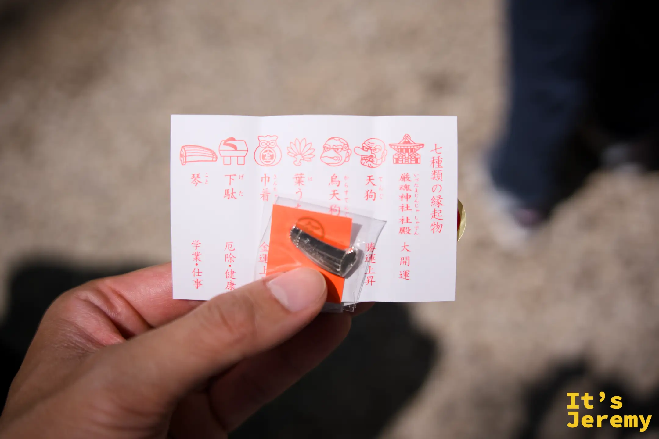 手持厳魂神社七種縁起物的說明卡片，七種圖案分別對應琴、下駄、巾着、葉、烏天狗、天狗、神社殿，旁邊有一個橘色小吊飾