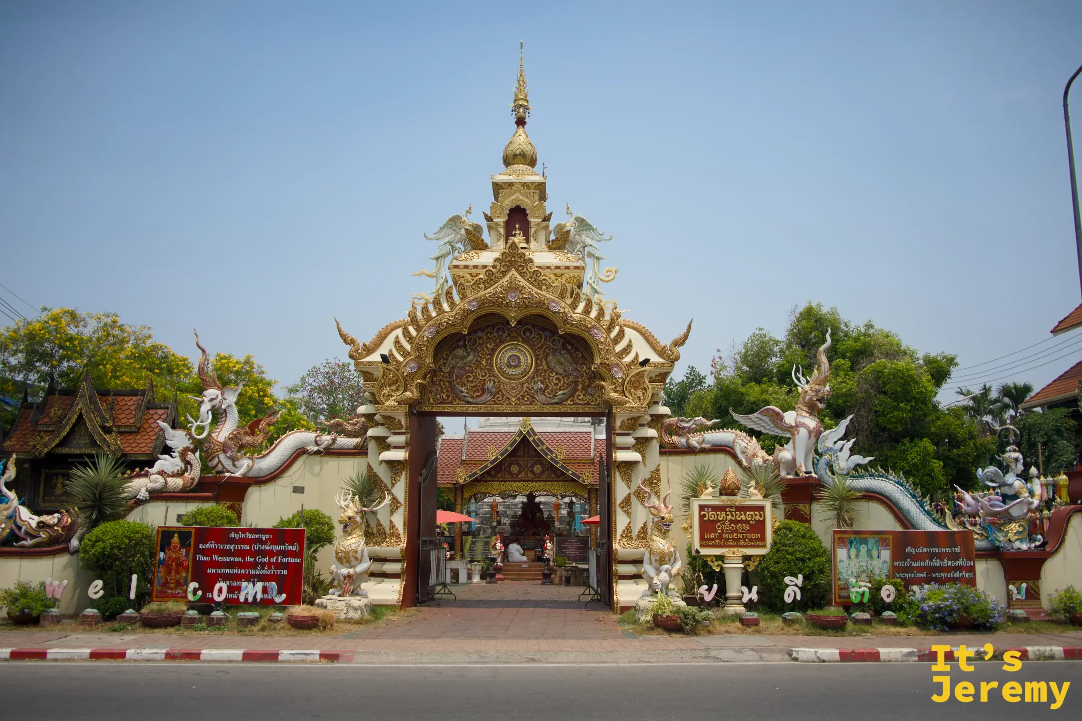 從帕辛路望向 Wat Muentoom 大門，金雕拱頂、那伽蛇身與守護神獸左右相對，門內隱約可見白色古塔