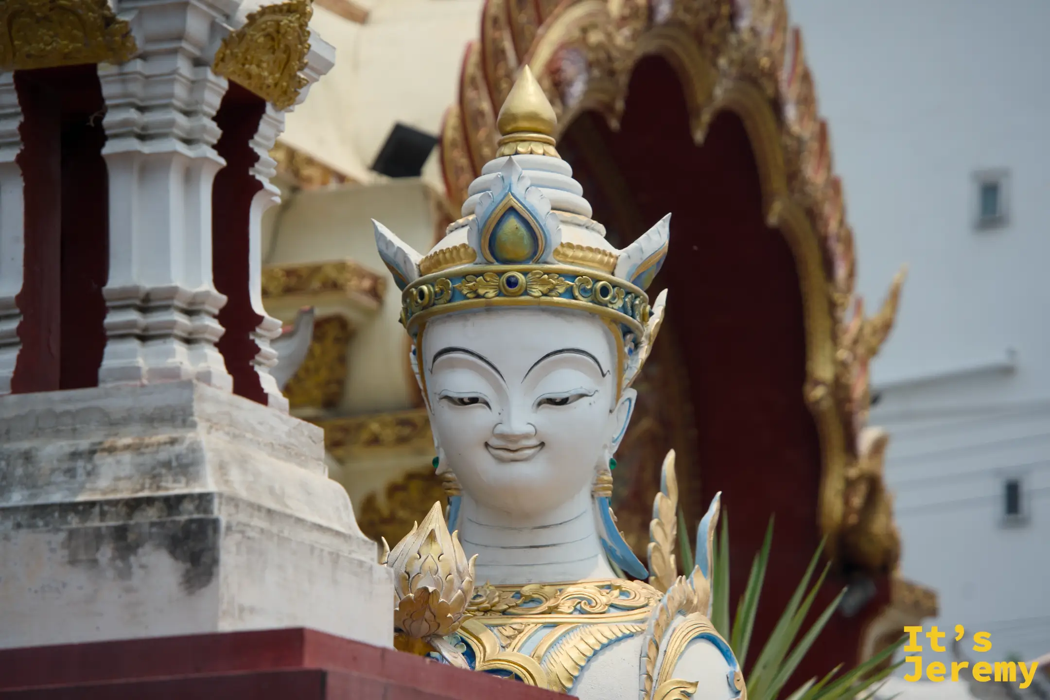 白色金冠的提婆達近景，眼神平靜，藍色瞳仁，後方可見寺廟建築的紅色拱門