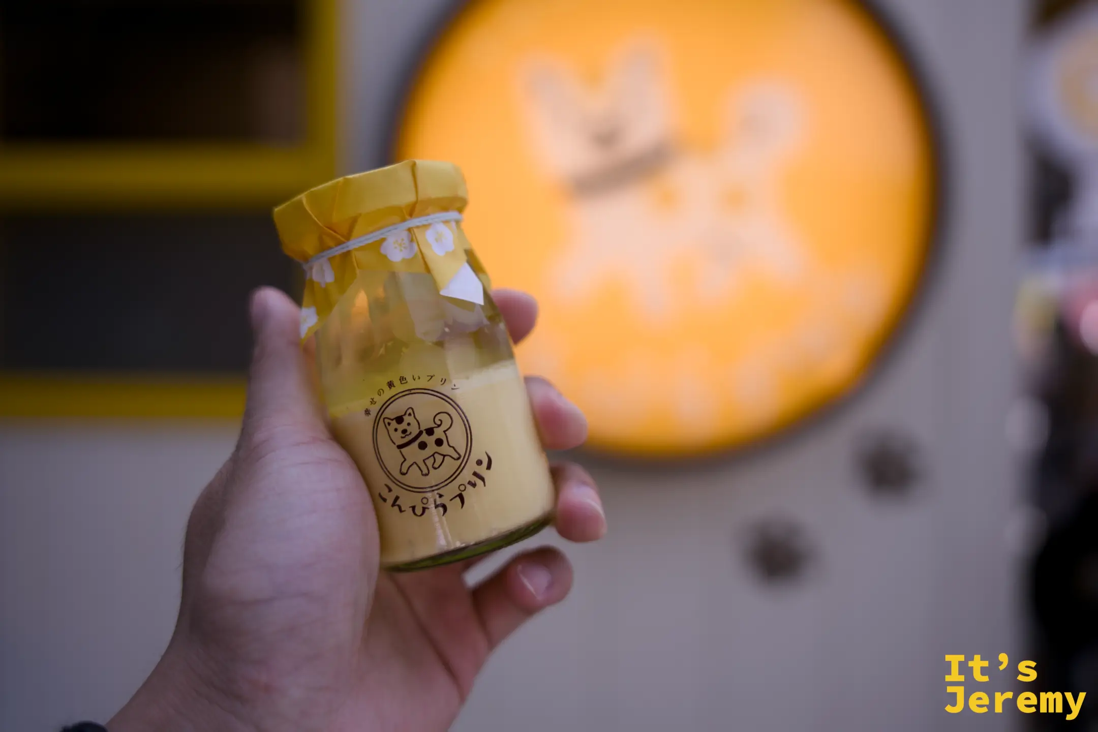 手持こんぴらプリン黃色布丁瓶，背後是橘色圓形招牌的模糊剪影，光線從後方透出