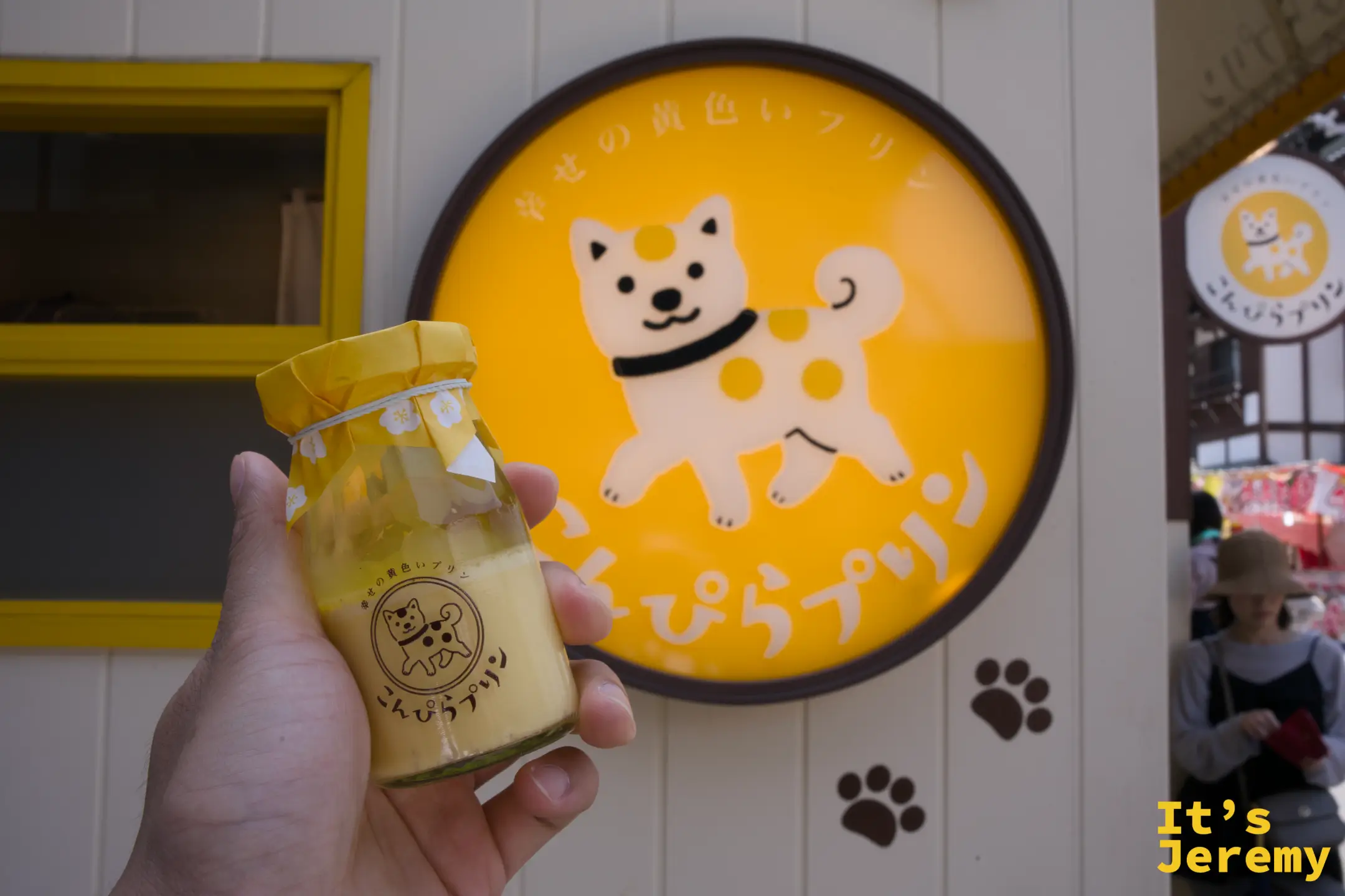 手持こんぴらプリン布丁瓶在店門口，背後是清晰的橘色大圓招牌，柴犬側面造型的圖案完整呈現