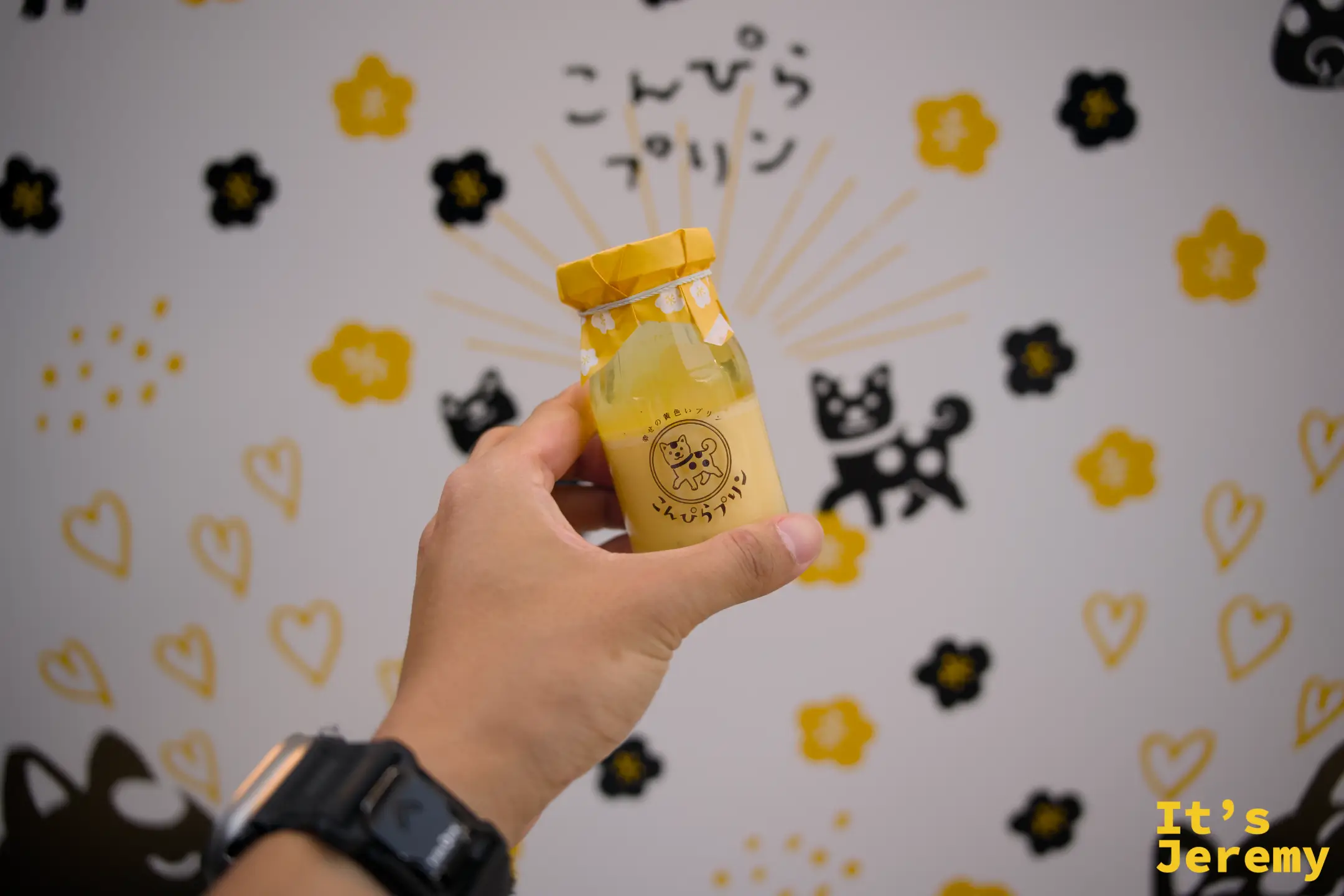 手持こんぴらプリン布丁瓶，背景是白色牆面上的黑黃柴犬花紋壁畫，店名字樣在上方