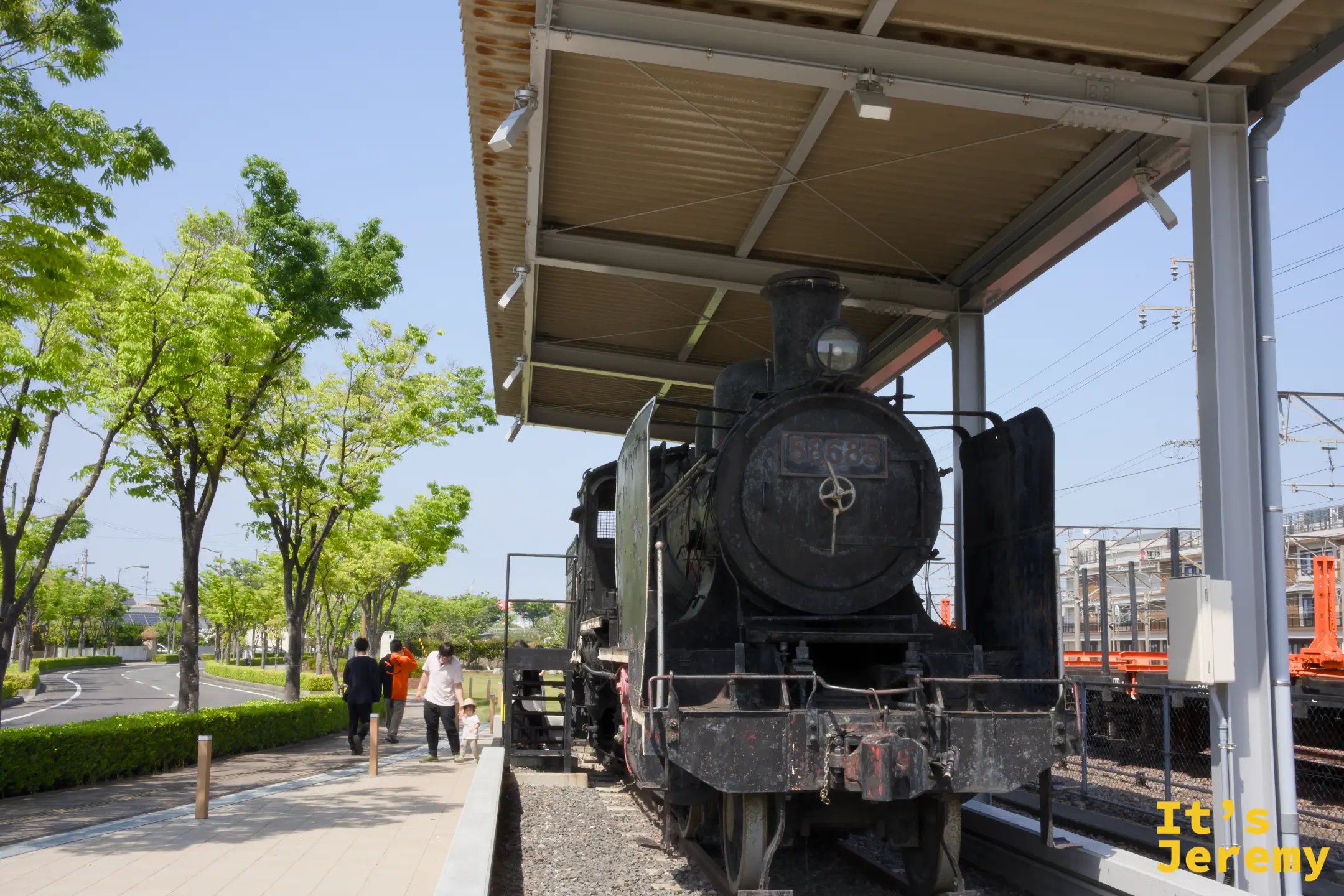 8620 型蒸汽機車正面仰視，棚架後方是藍天和綠樹，遠處有行人走過