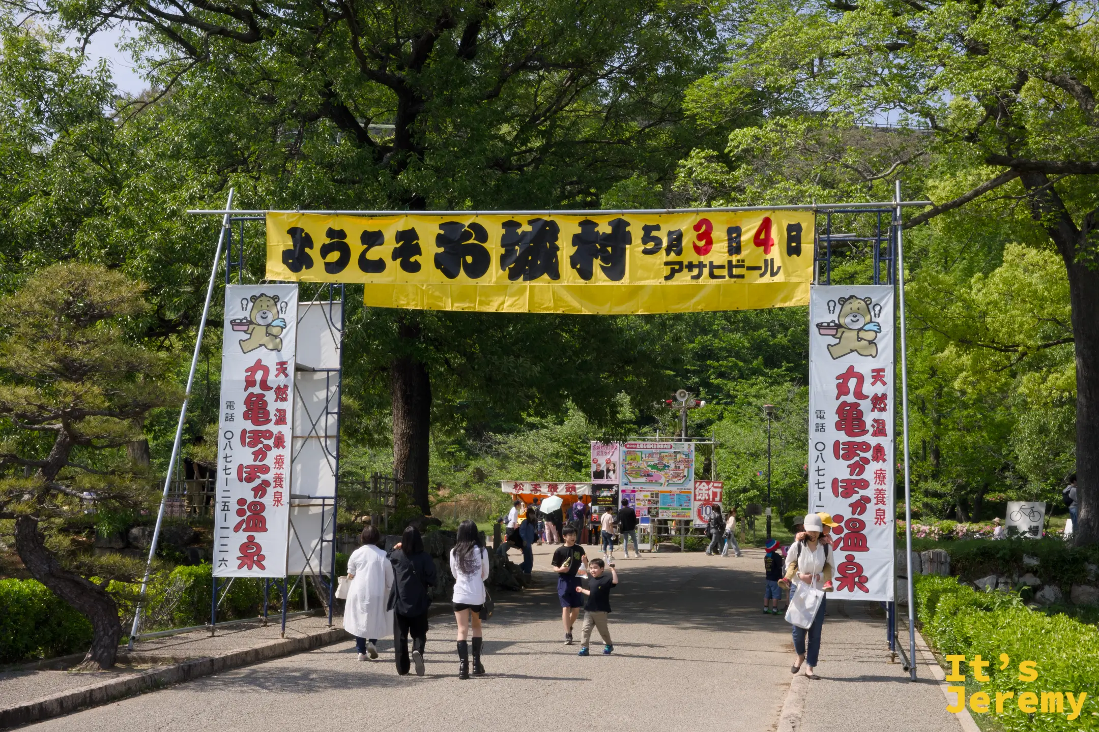 入口拱門掛著「ようこそお城村」的黃色橫幅，杉樹夾道，遊客陸續進入