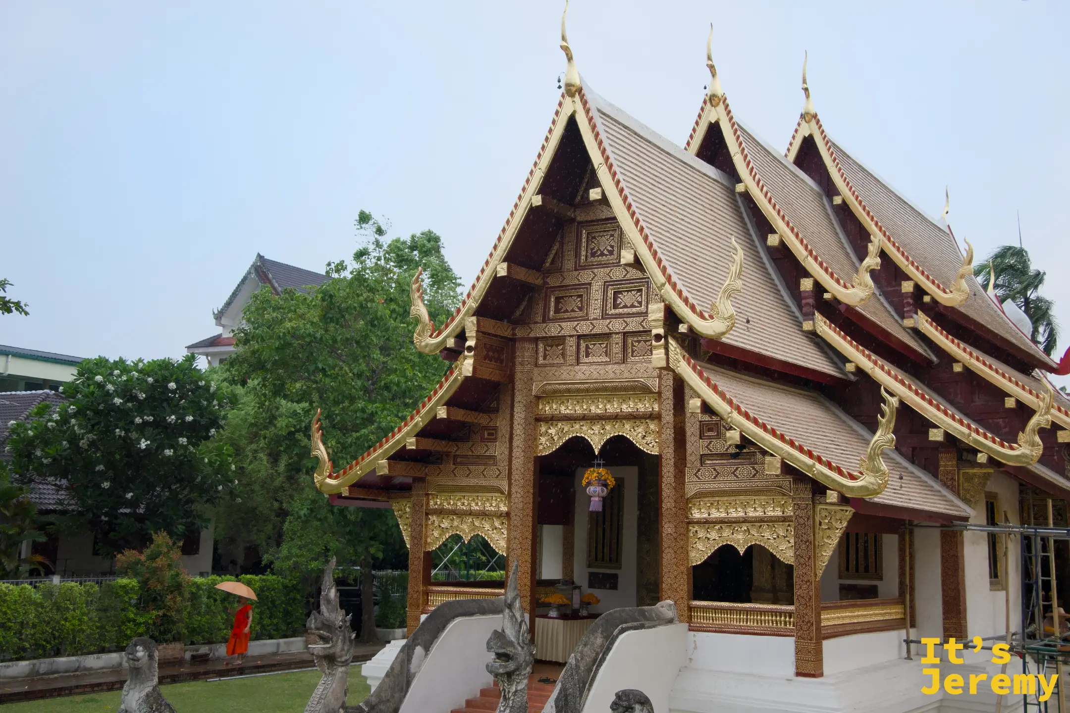 偏殿建築，多重蘭納屋簷層疊，一名橘衣僧侶撐傘走過，雨後寂靜