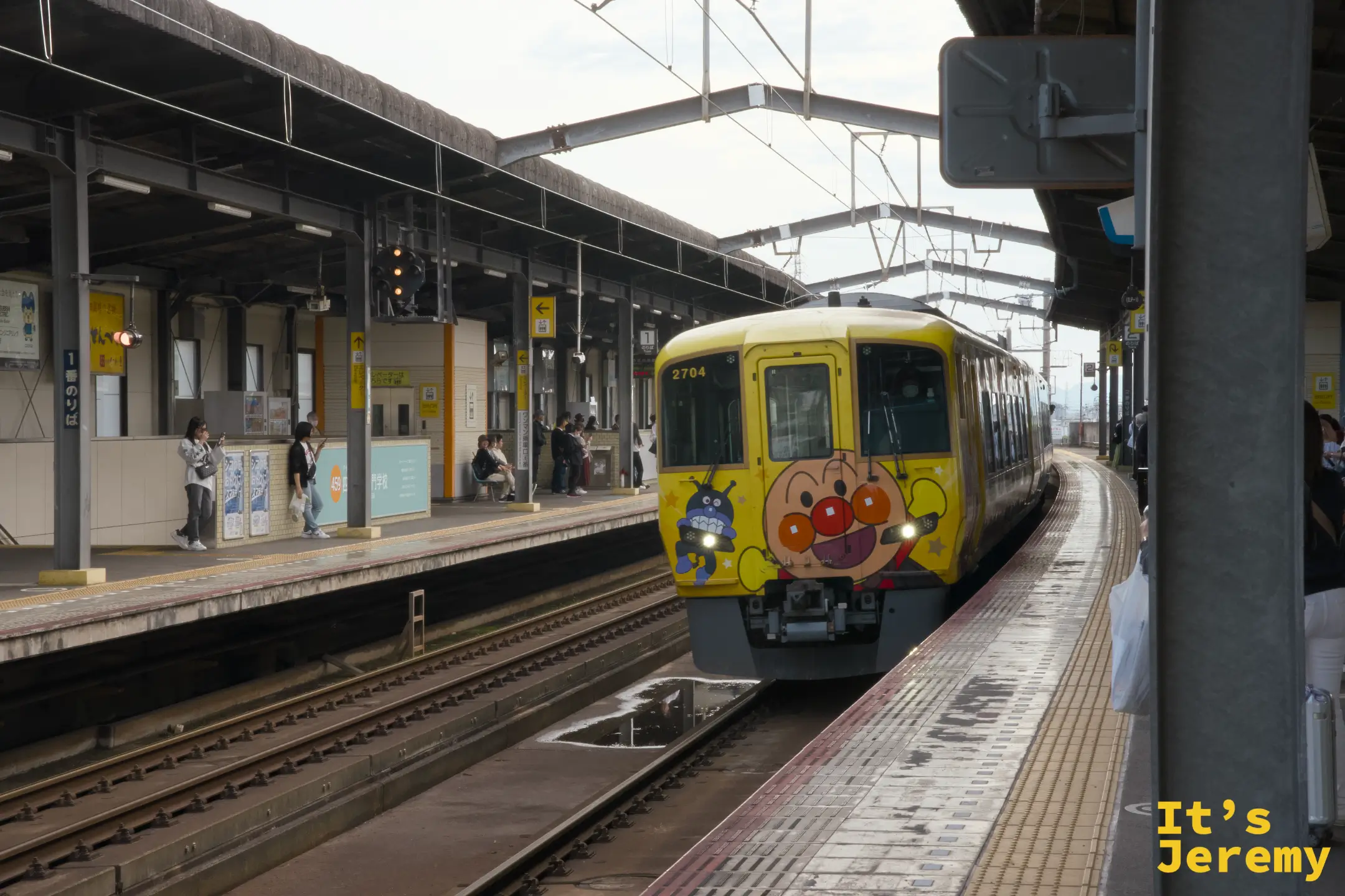 黃色麵包超人列車（2704）從遠方駛入，月台兩側有乘客等候，車頭麵包超人圖案鮮豔