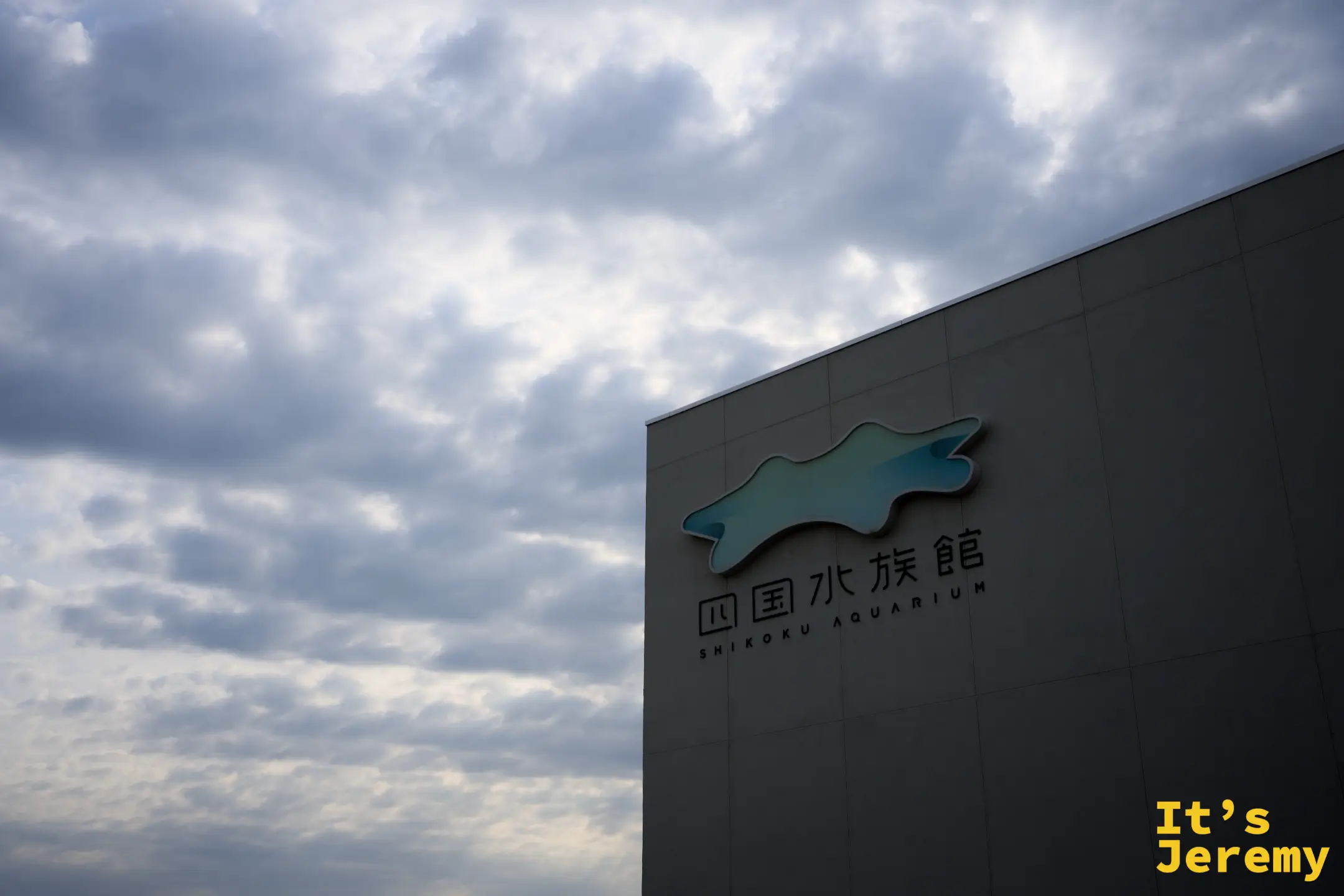四國水族館外觀，白色低矮建築側牆掛著魚形 LOGO，背景是多雲的天空