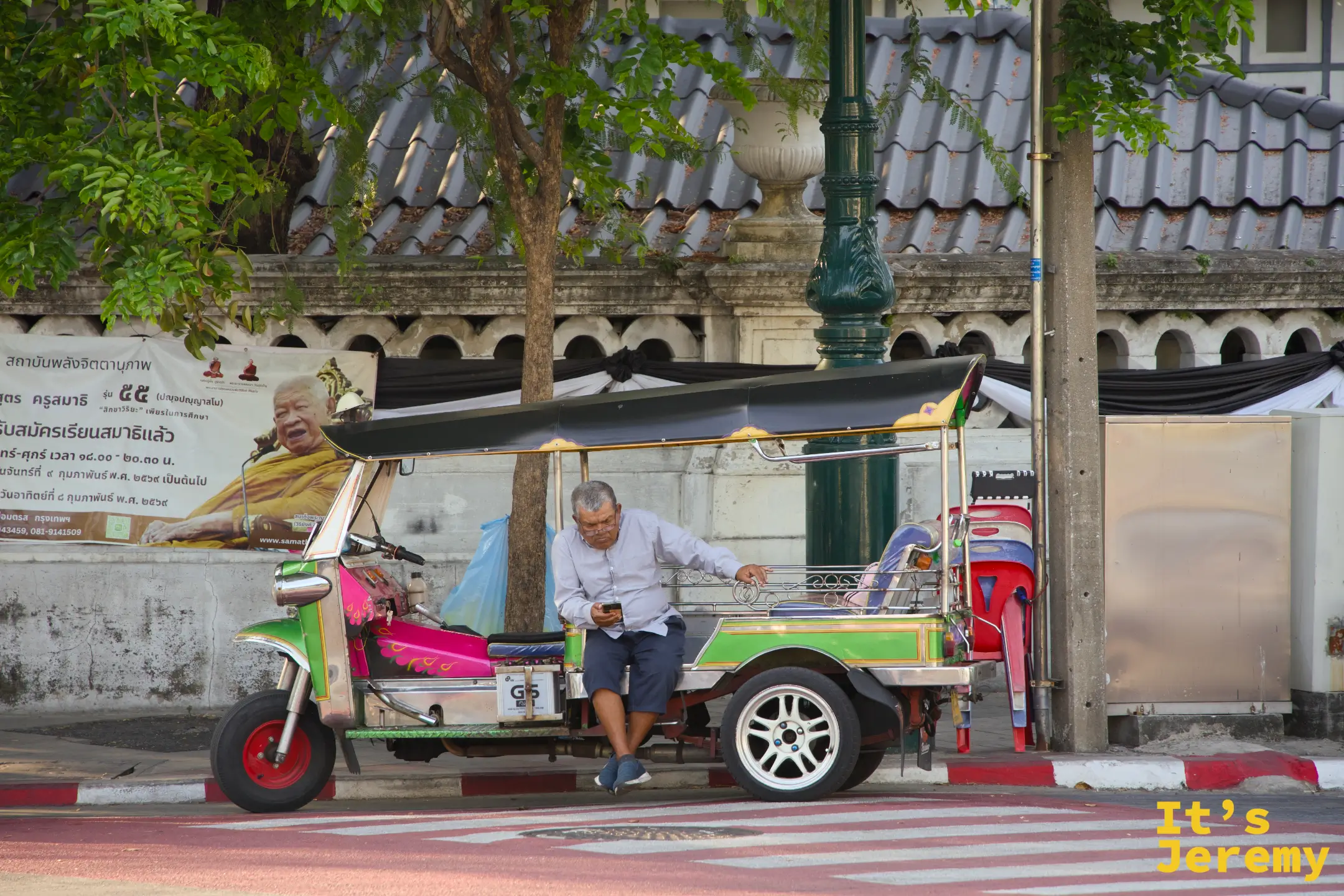一輛停在運河邊的紅藍 tuk-tuk、司機坐在駕駛座上滑手機，後座掛著一張高僧海報