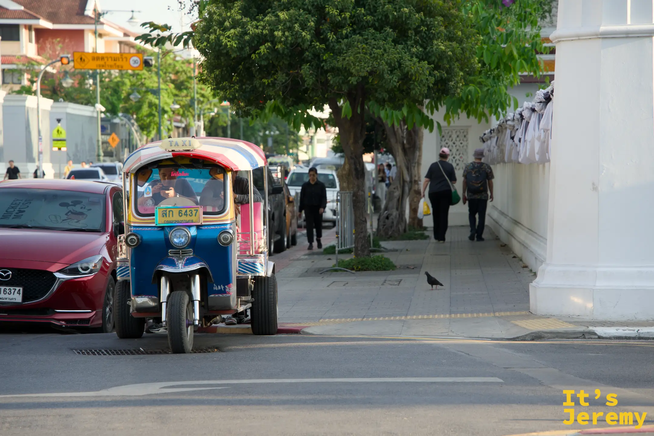 一輛藍色 tuk-tuk「ส 6437 TAXI」從紅色 Mazda 旁邊開過、遠處走著一對老夫妻——通往 Tha Tien 碼頭最後 200 公尺