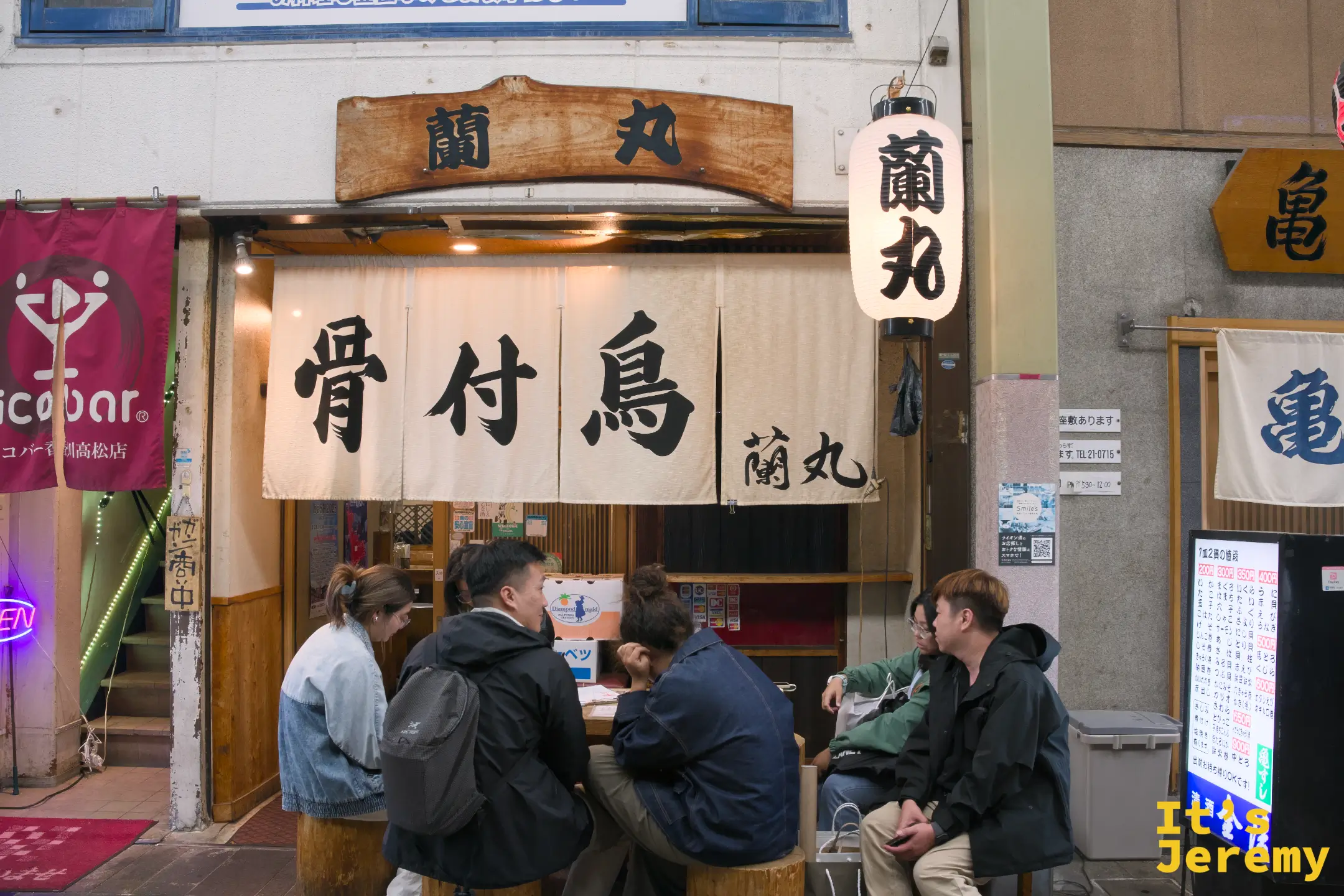 蘭丸商店街店面正面全景，骨付鳥大字暖簾與「蘭丸」木招牌，客人在外等位