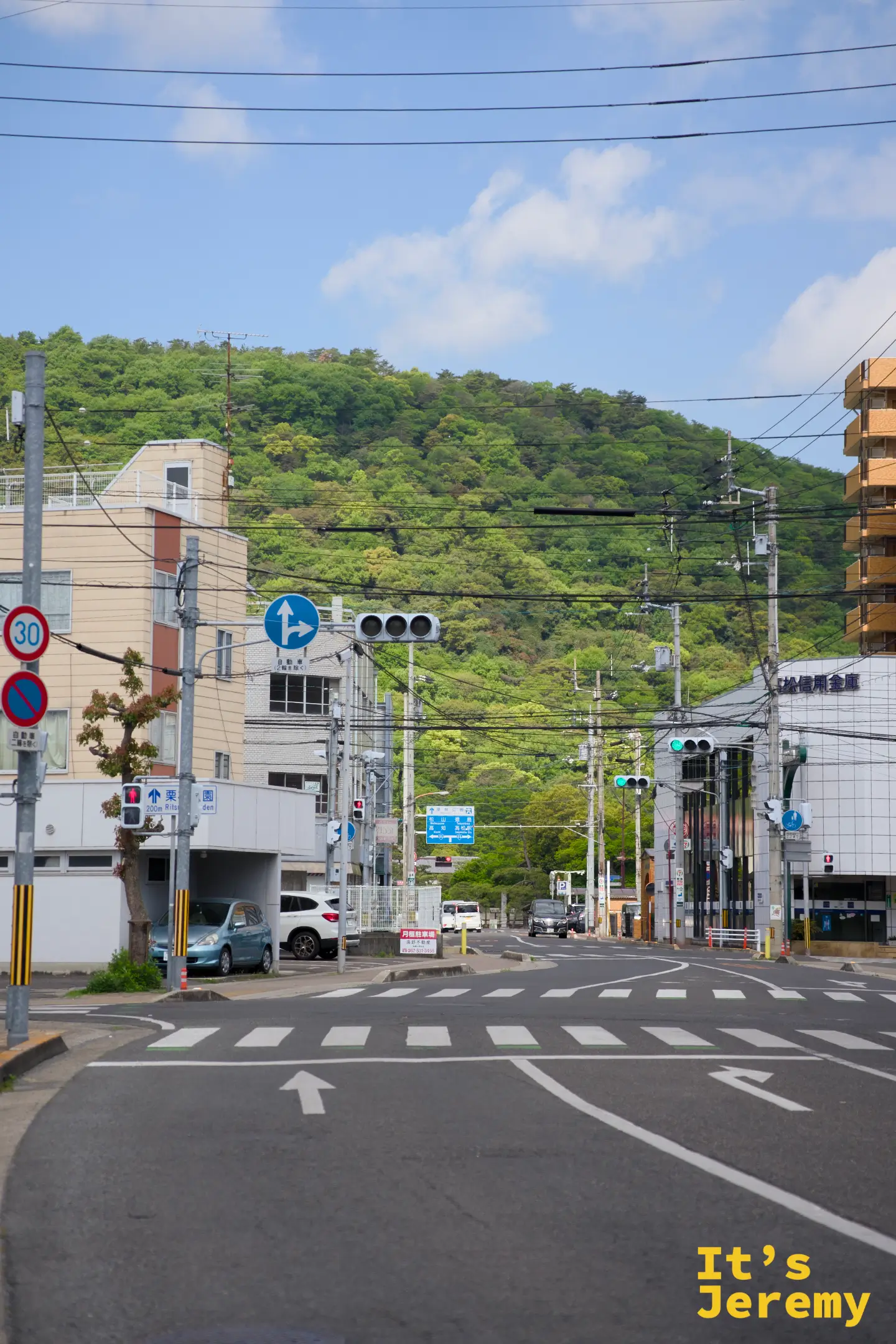 高松市區街道，前方一排建築物與商店，背後是一道緩坡起伏的綠色丘陵