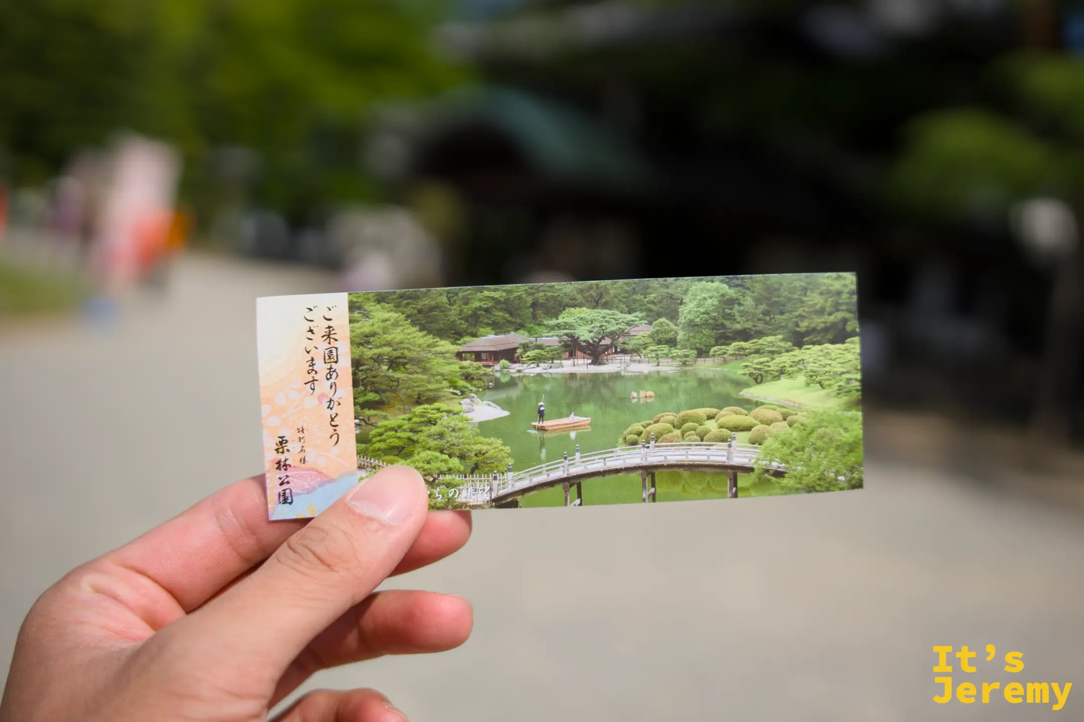 手持栗林公園入場券，背景是園內的傳統建築與綠樹