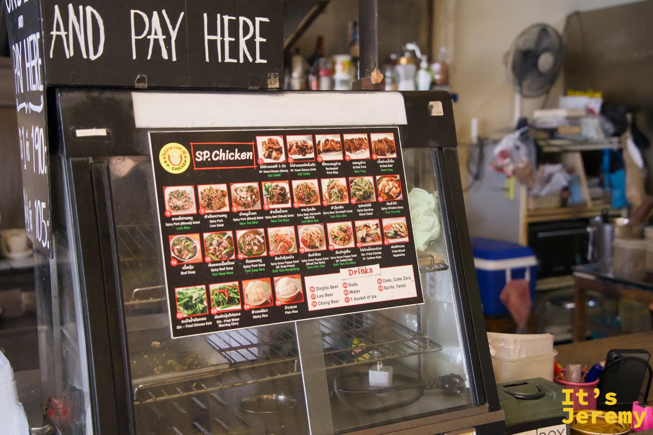 店內結帳台，玻璃冷藏櫃上貼著彩色圖文菜單，黑板寫著 AND PAY HERE
