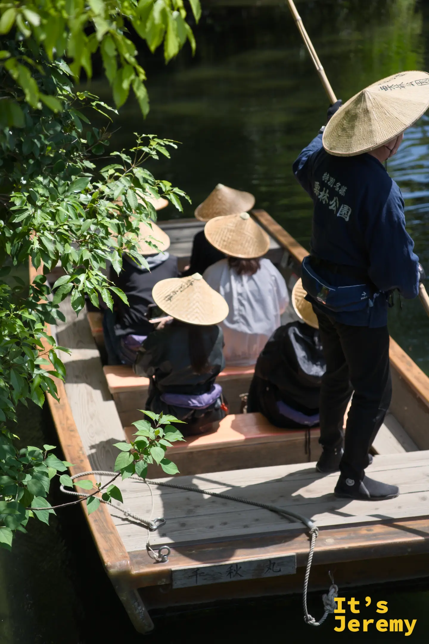 船伕和乘客在傳統和船上的畫面，船伕身穿深藍色栗林公園制服、戴著斗笠撐篙，乘客同樣戴著斗笠坐於船中