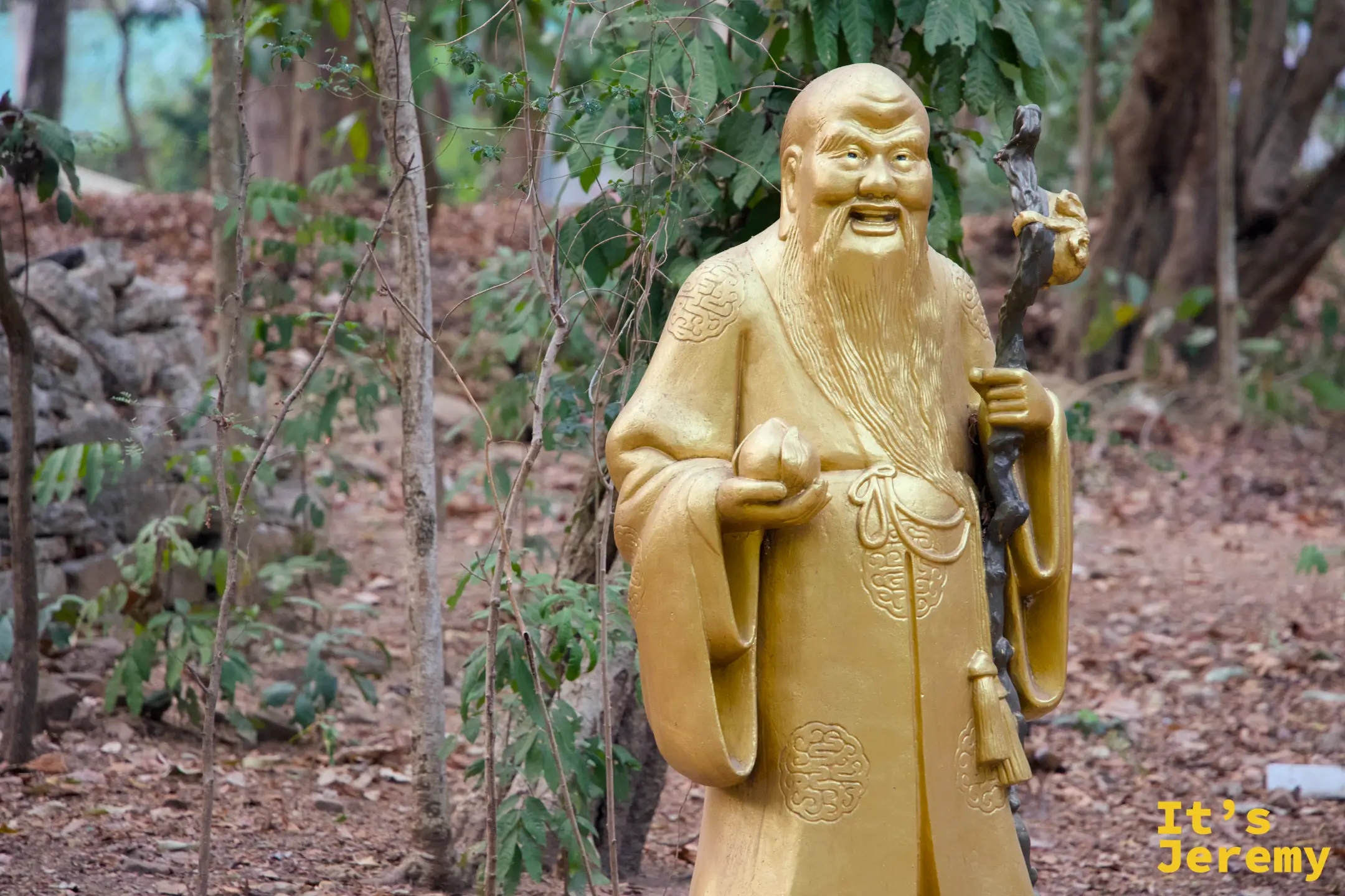 中式老者雕像，持杖微笑，背靠綠樹