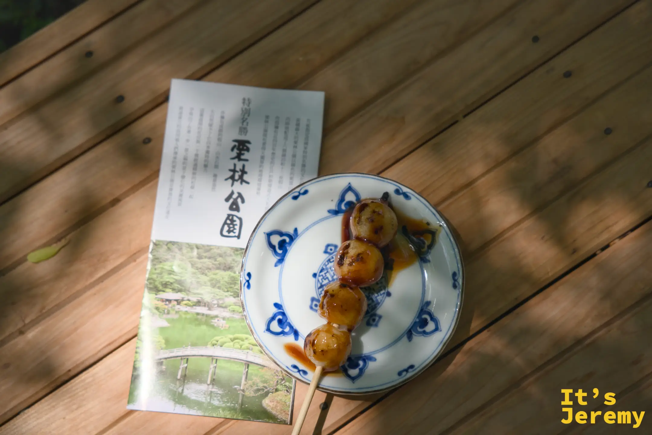 木造桌面上，一串糰子放在青花瓷小盤上，旁邊是攤開的栗林公園導覽手冊，手冊封面是偃月橋的照片