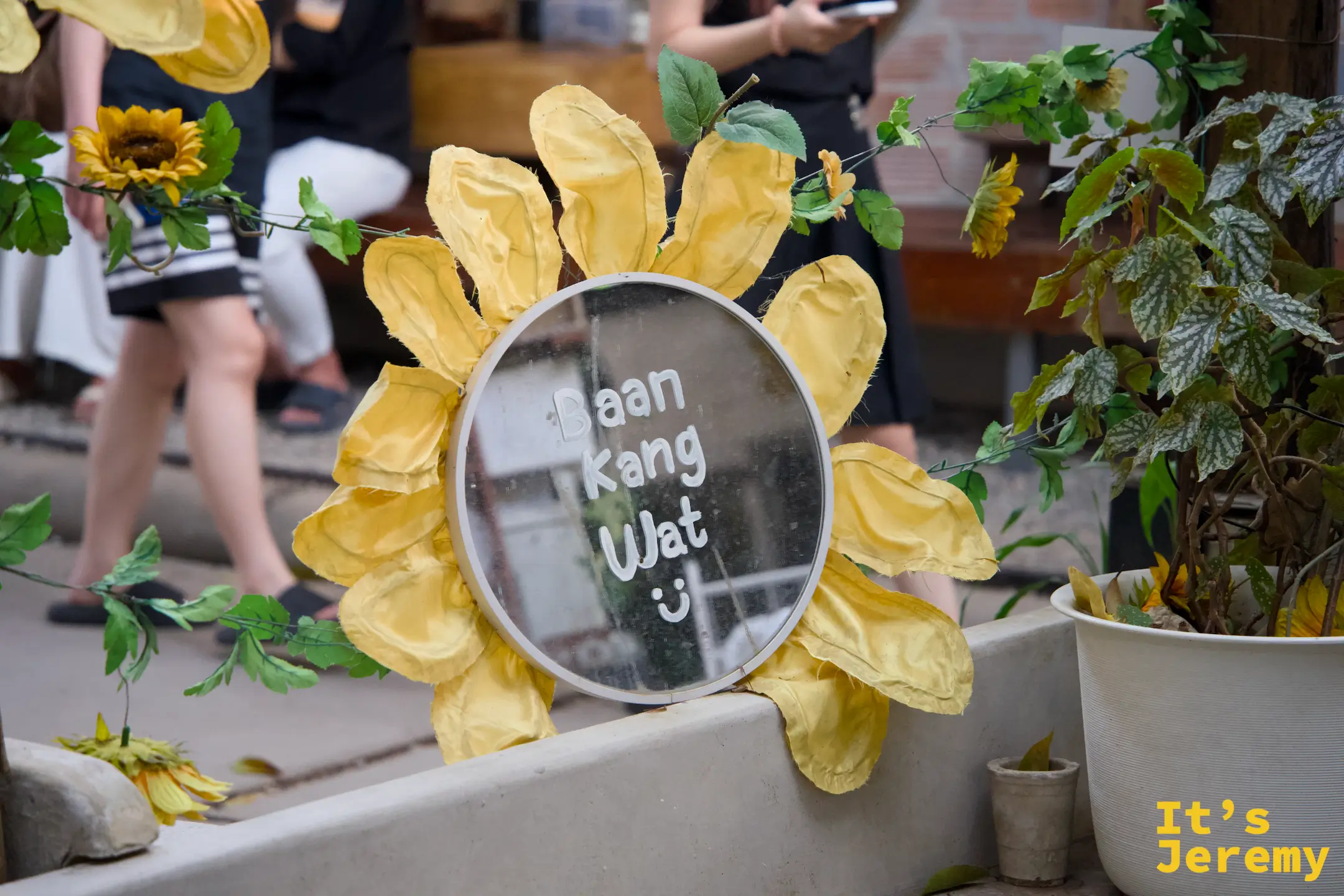 向日葵花框圓鏡，中央鏡面上寫著「Baan Kang Wat :)」，黃色布料花瓣圍繞，旁邊有盆栽和綠葉點綴