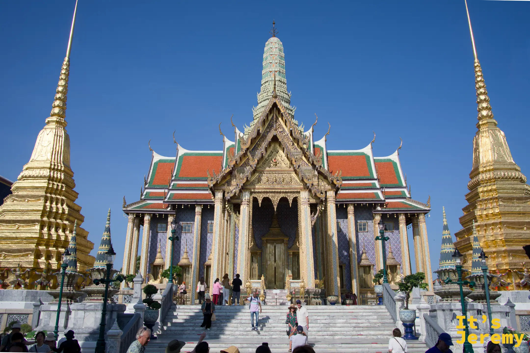 玉佛寺主殿正面全景，左右兩座金色 Chedi 與遊客上下台階