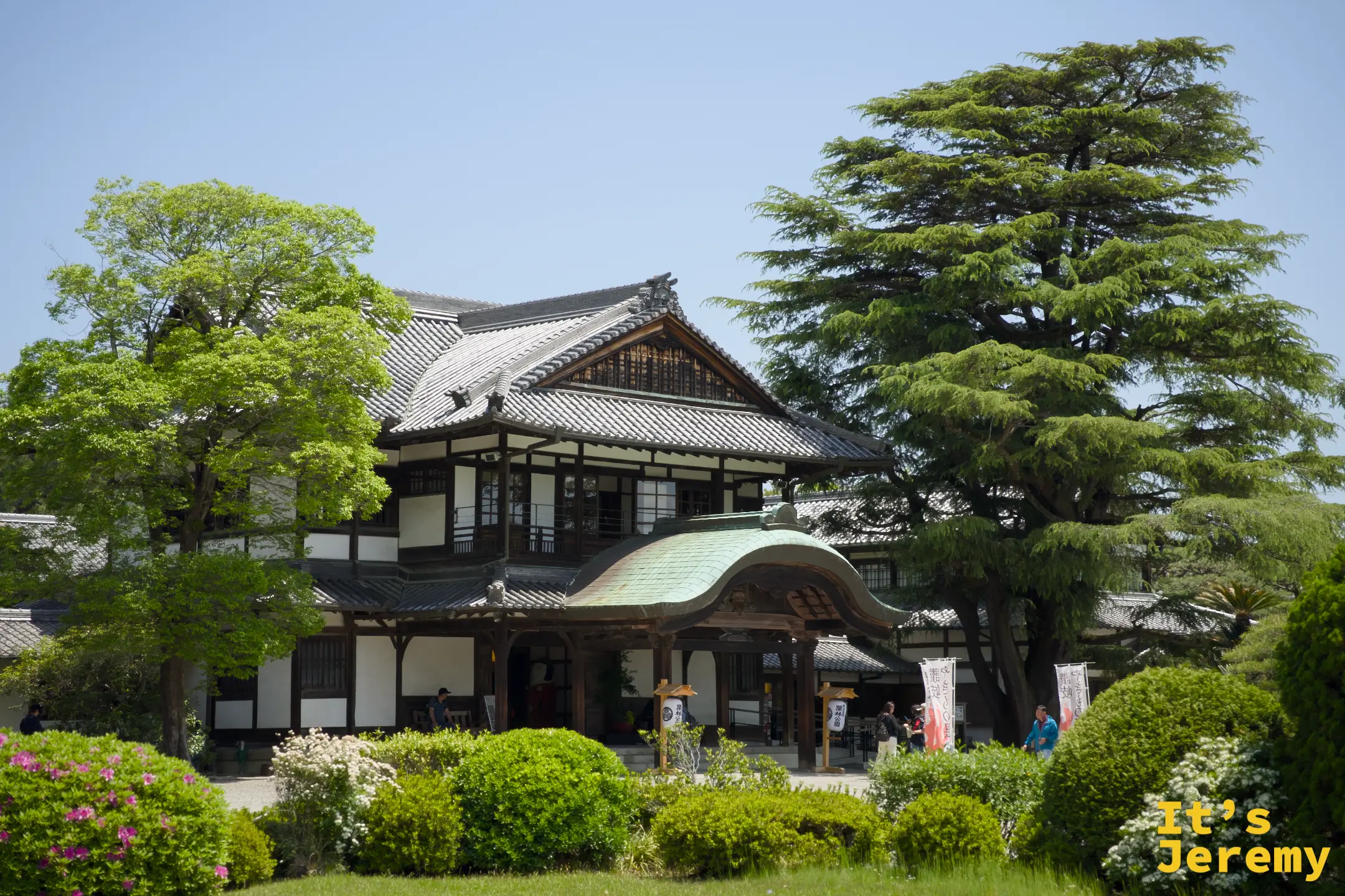 工商獎勵館正面，兩層樓傳統木造建築配上青瓦屋頂，四周圍繞著松樹和闊葉樹，前方有杜鵑花叢