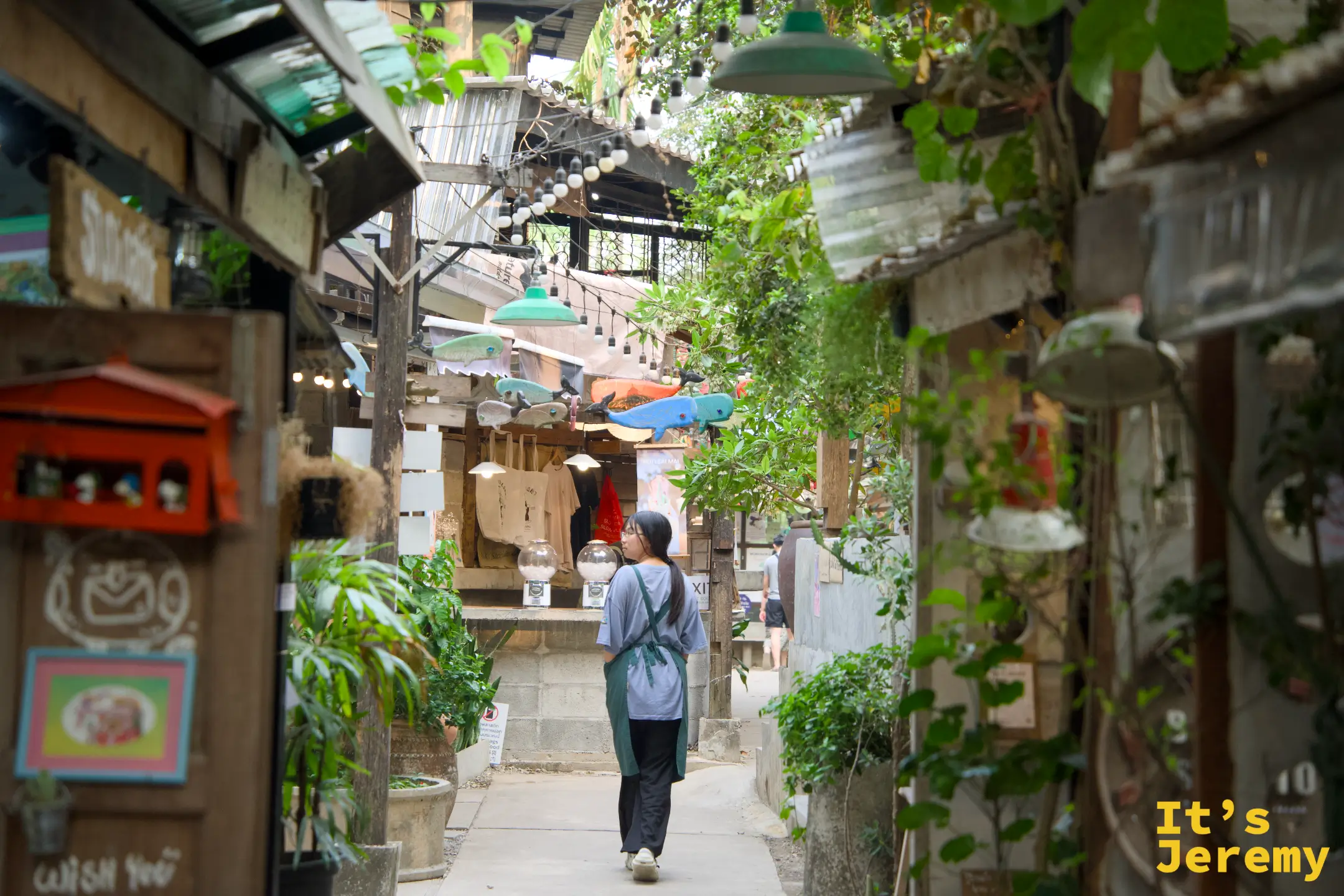 穿圍裙的工作人員走在 Baan Kang Wat 的巷弄間，兩側是藤蔓、植物與各家獨立小店