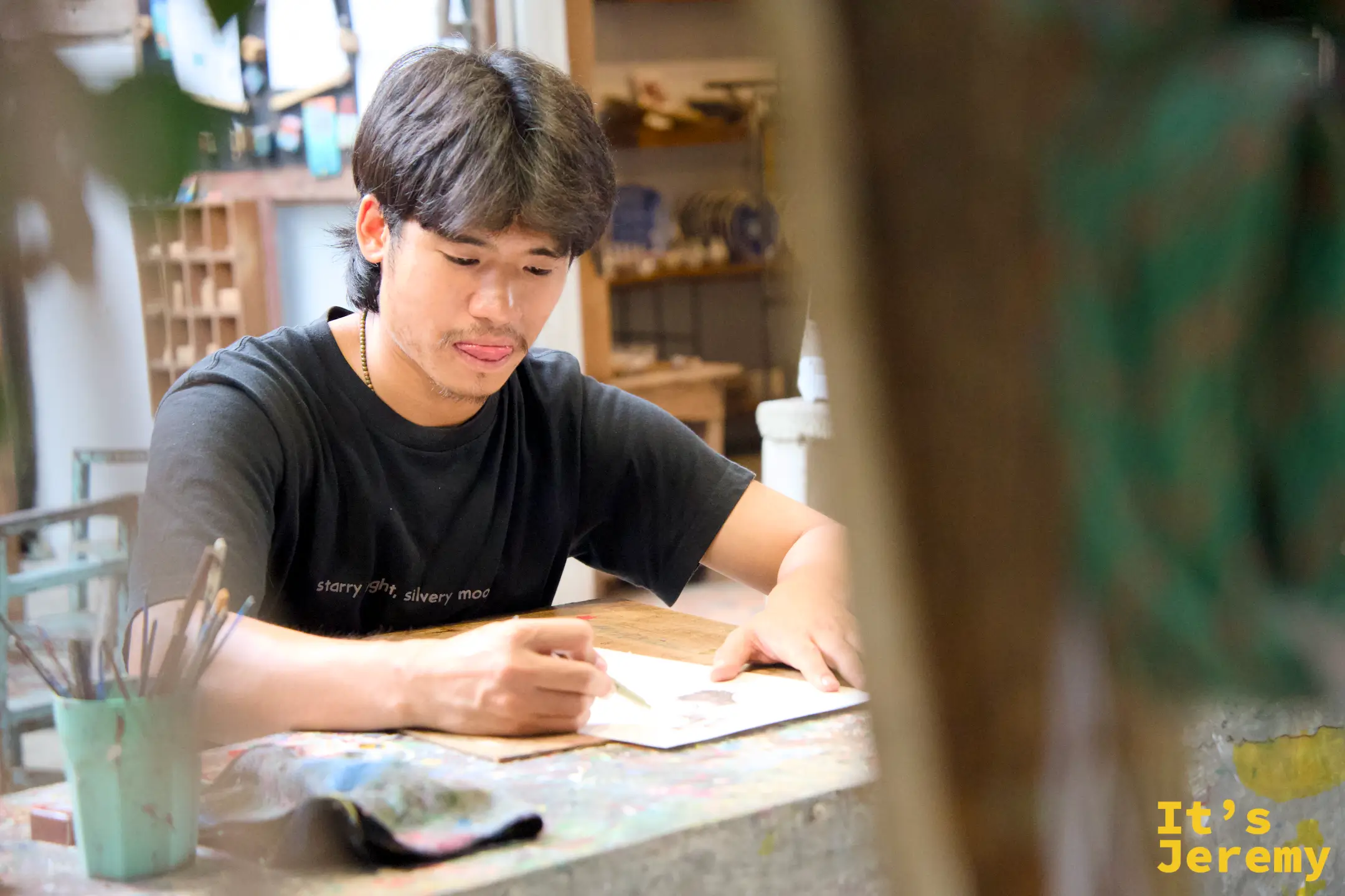 年輕男性在工作室內低頭創作，桌上有顏料調色盤和畫筆，後方是陶瓷作品展示架