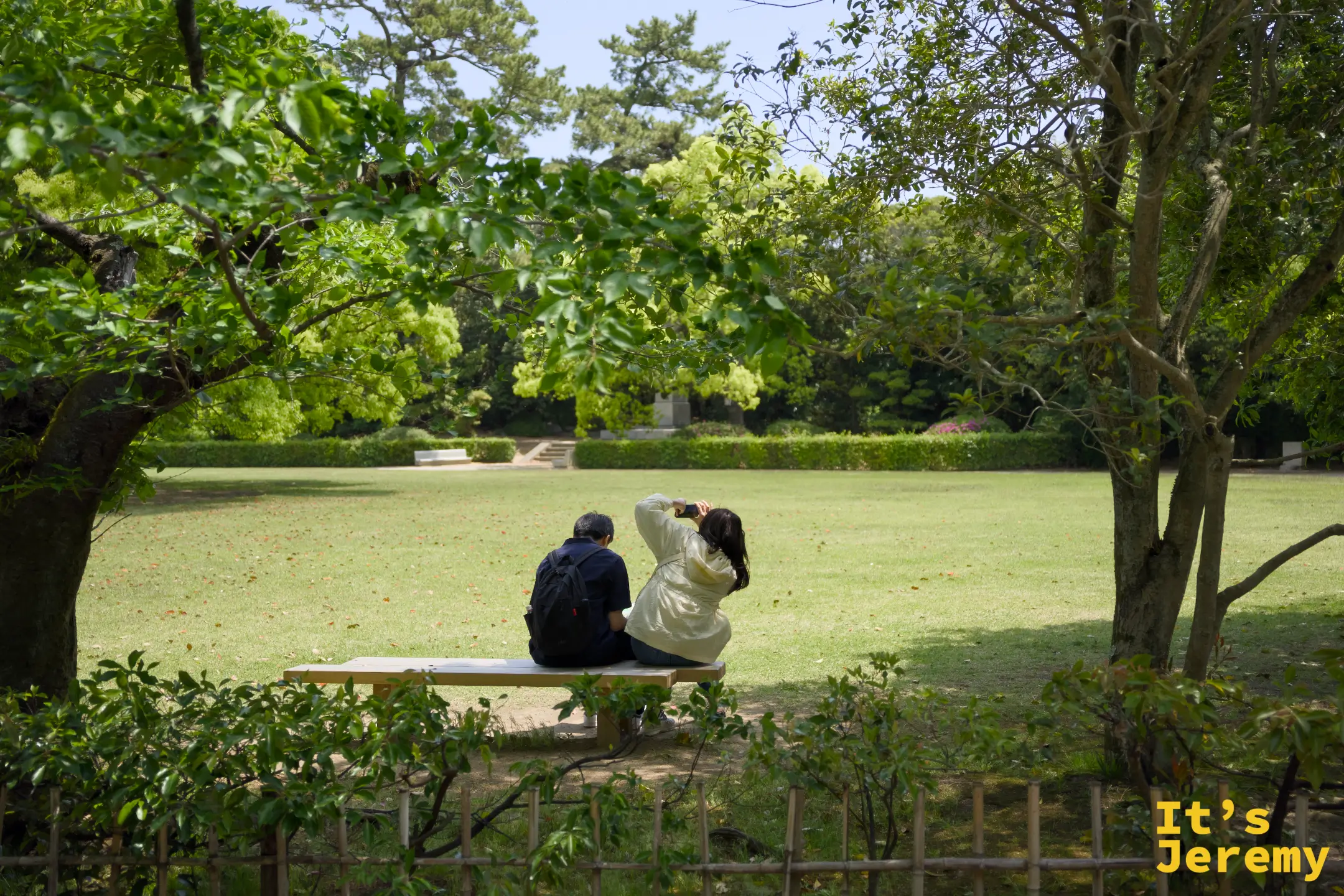 一對夫婦背對鏡頭坐在長椅上，面向開闊草地和遠處樹林，坐姿安靜，像是在曬太陽