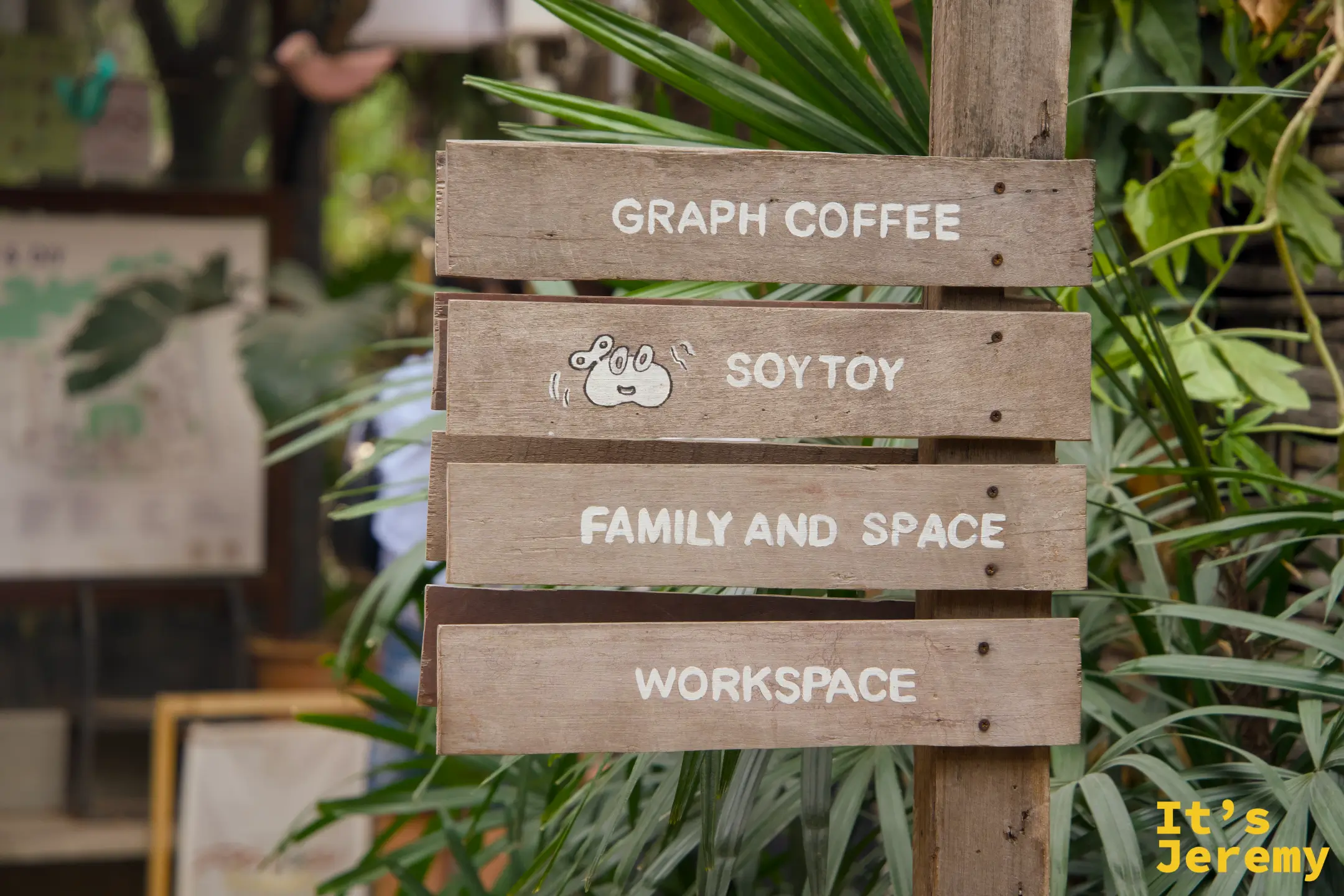 木製指示牌「GRAPH COFFEE / SOY TOY / FAMILY AND SPACE / WORKSPACE」，被綠色植物包圍