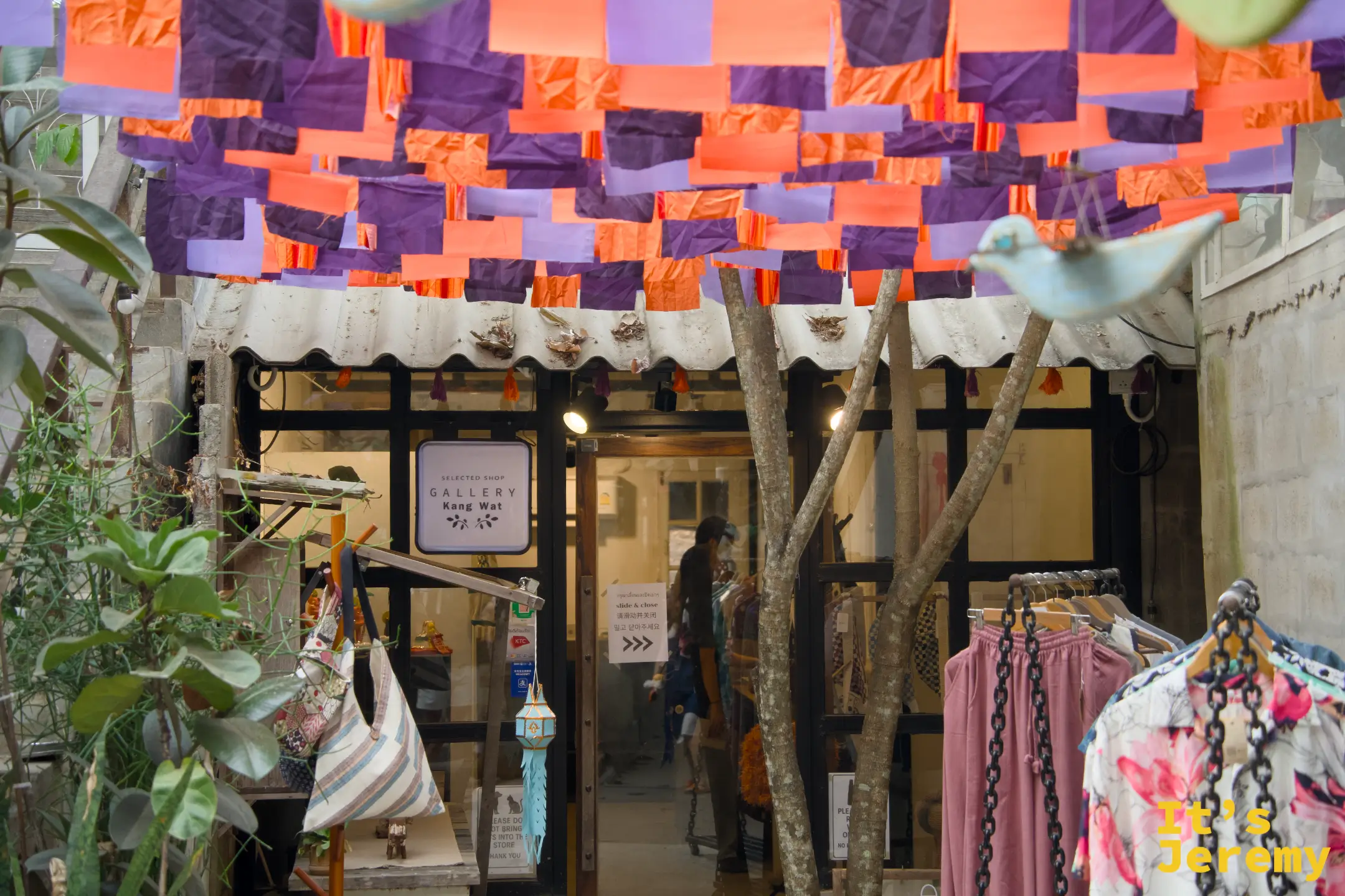 Gallery Kang Wat 選品店外觀，天花板懸掛橘紫布幔，店門口有衣架展示服裝，樹枝裝飾立在入口