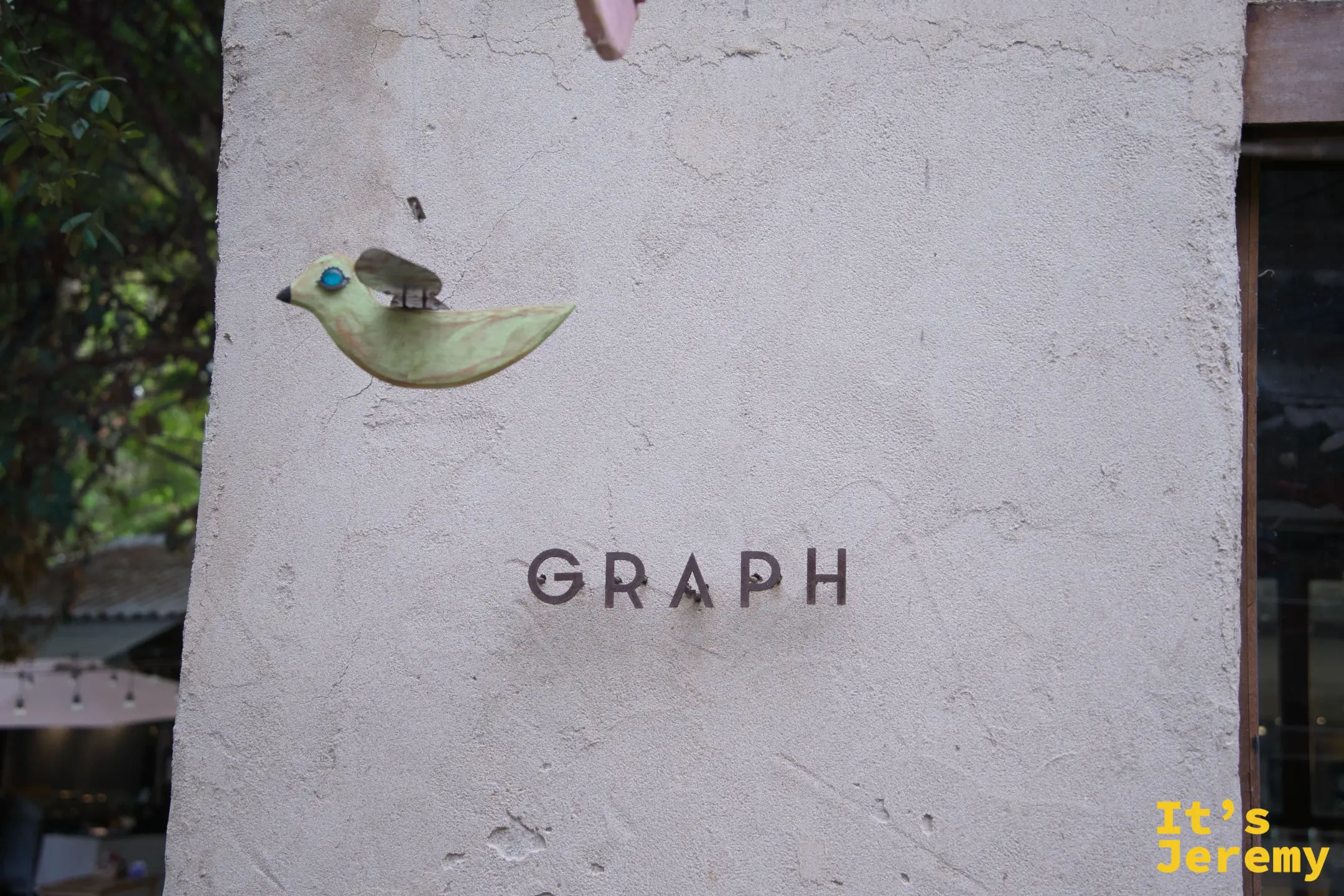 水泥外牆近拍，左側有一隻木雕小鳥裝飾，右側以簡約字體刻著「GRAPH」，風格克制精緻