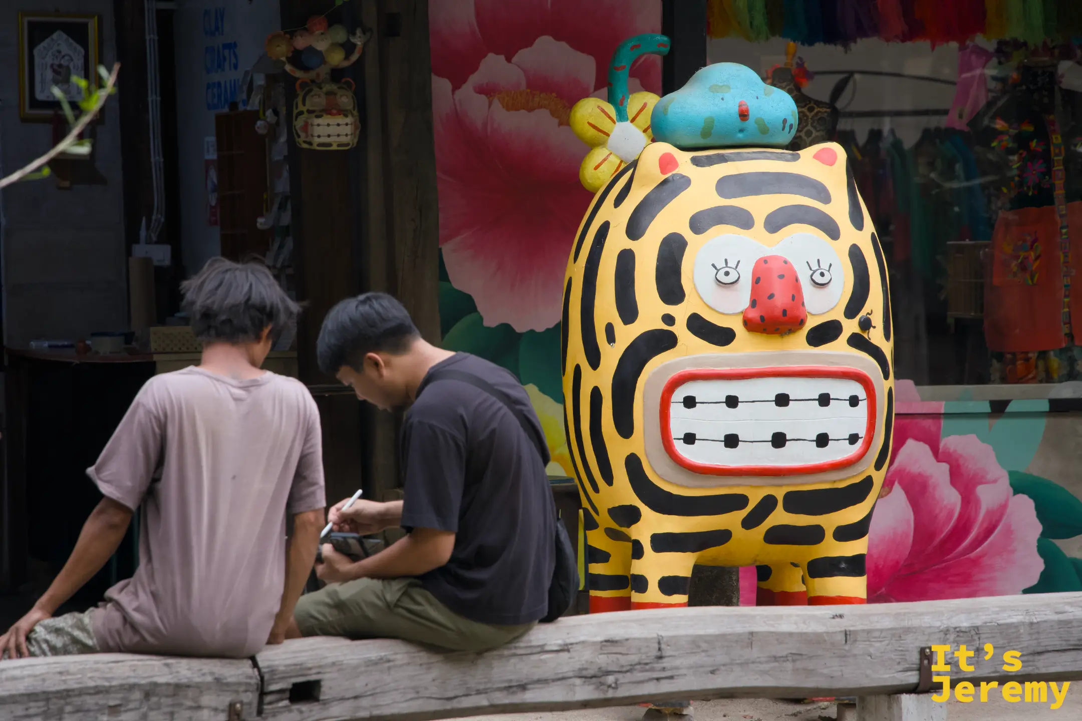 兩個男生坐在大型老虎吉祥物旁，神情悠閒，一人低頭看手機，旁邊可見彩色商品陳列
