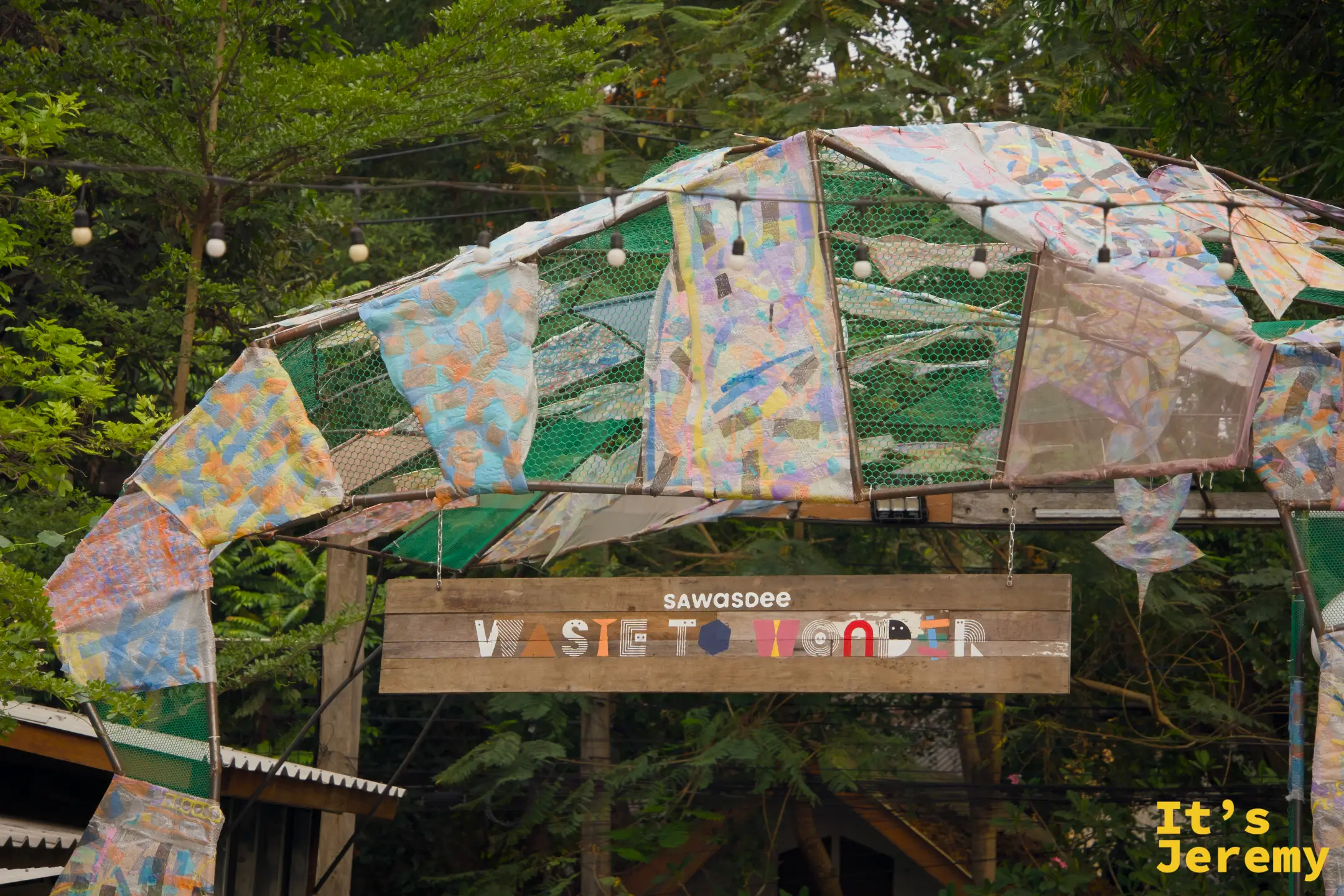 「sawasdee WASTE TO WONDER」廊架全景，由廢棄材料拼接成魚形弧頂，上方是彩色廢布帆，在綠樹背景下格外顯眼