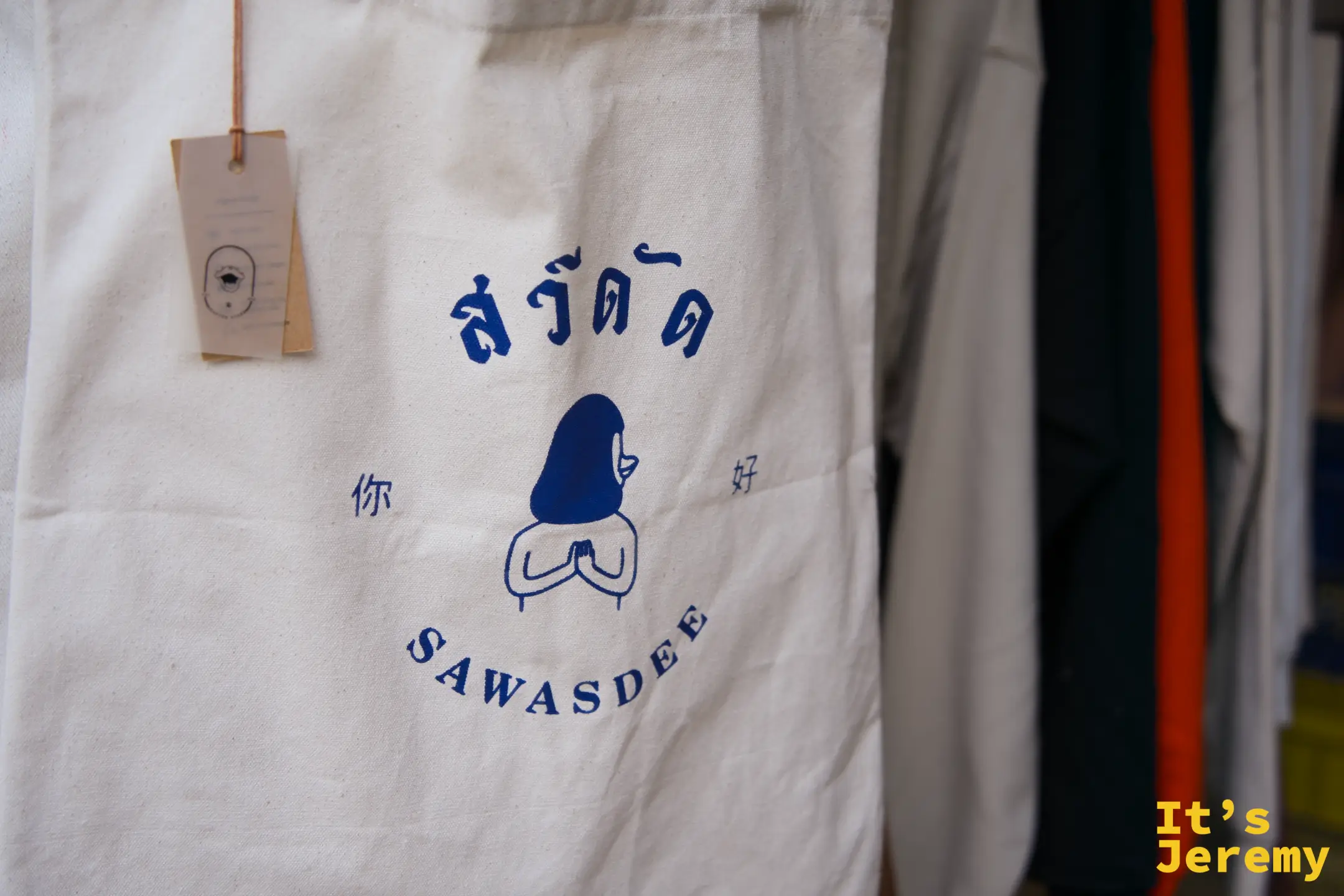 白色帆布托特包特寫，印著泰文「สวัสดี」、中文「你好」和「SAWASDEE」藍色合掌人偶圖案