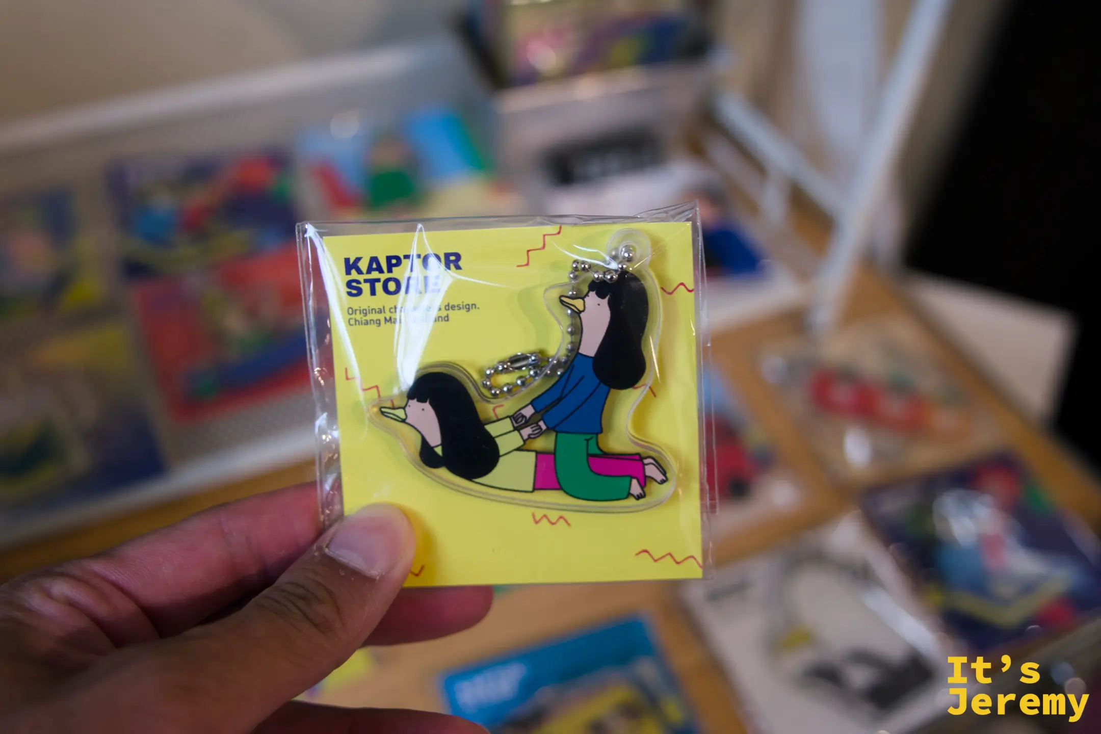 手持 Kaptor Store 鴨子角色壓克力鑰匙圈，黃色背景卡紙，後方可見更多插畫小商品