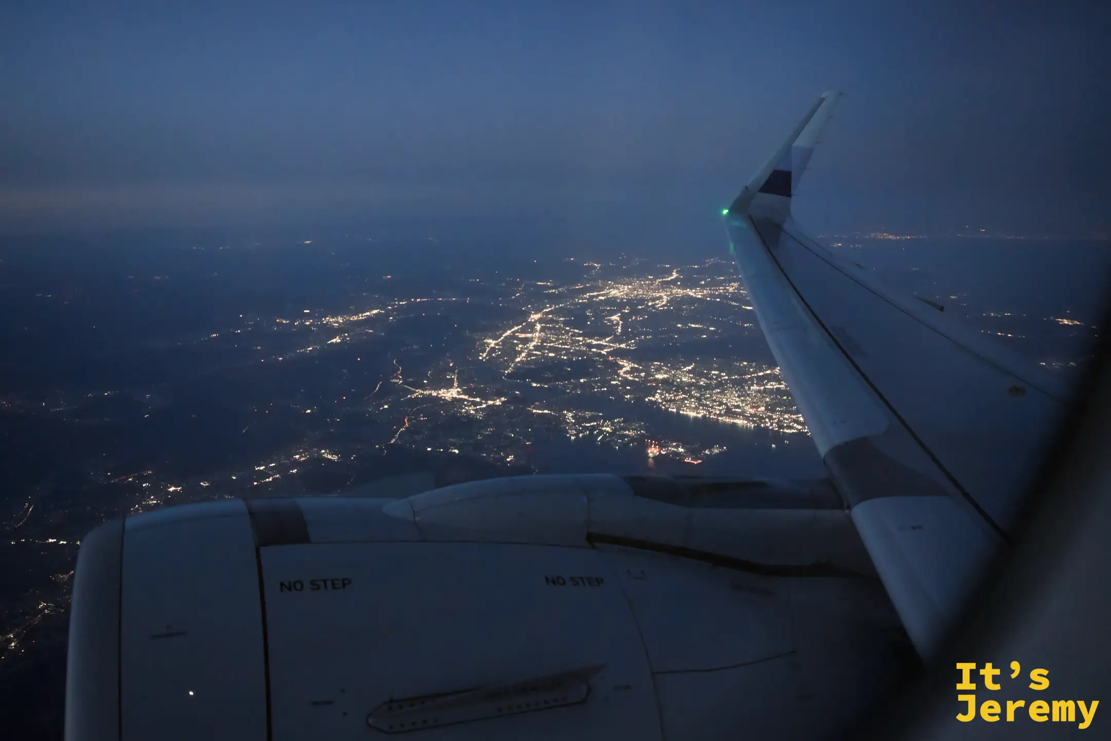 夜間從飛機窗口俯瞰，機翼前方是大片金黃色城市燈光網絡，深藍夜空與燈光地面之間截然劃分