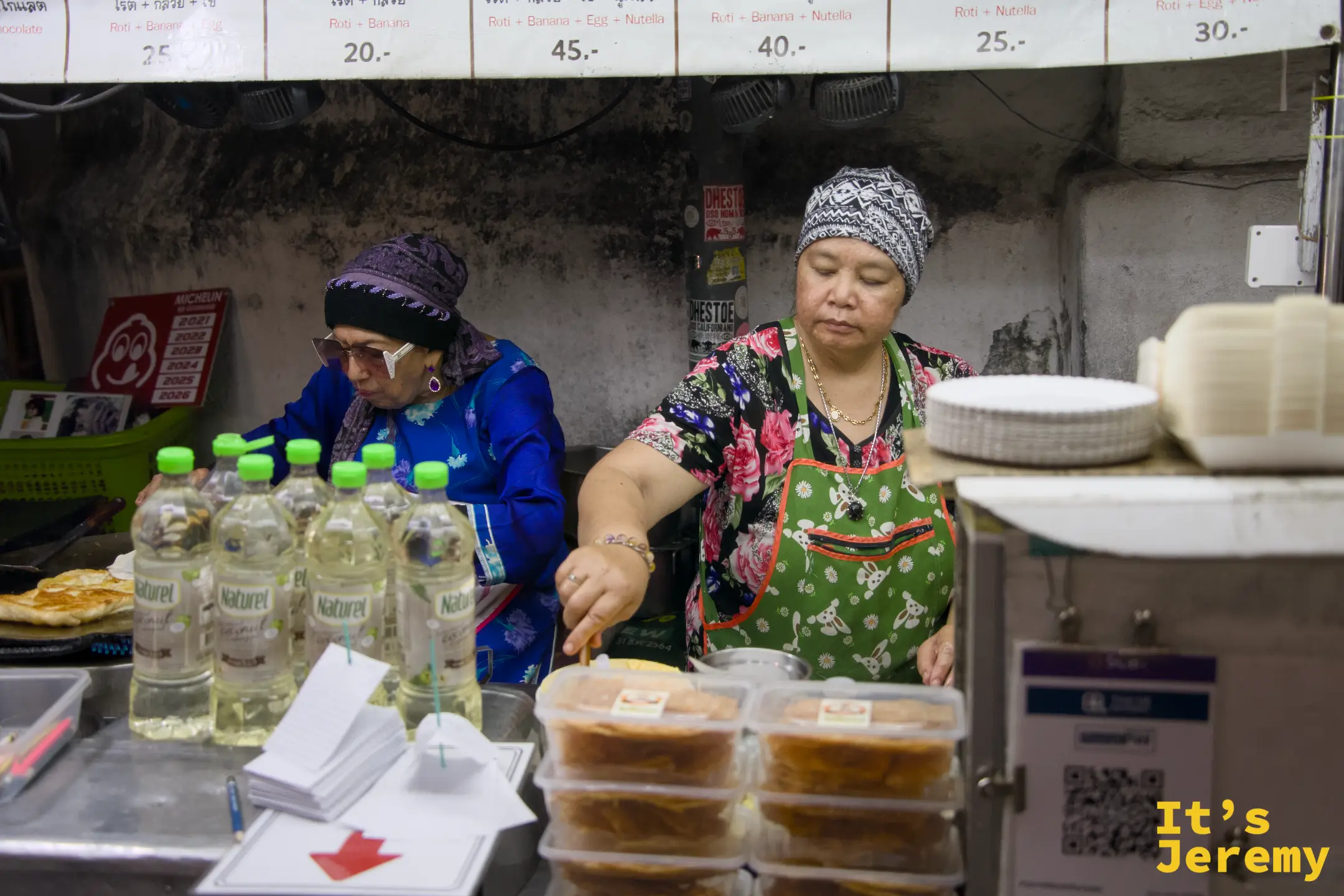 兩位掌攤的阿姨：左側戴頭巾的奶奶顧著鐵板，右側綠圍裙阿姨打理檯面，上方掛著價目表