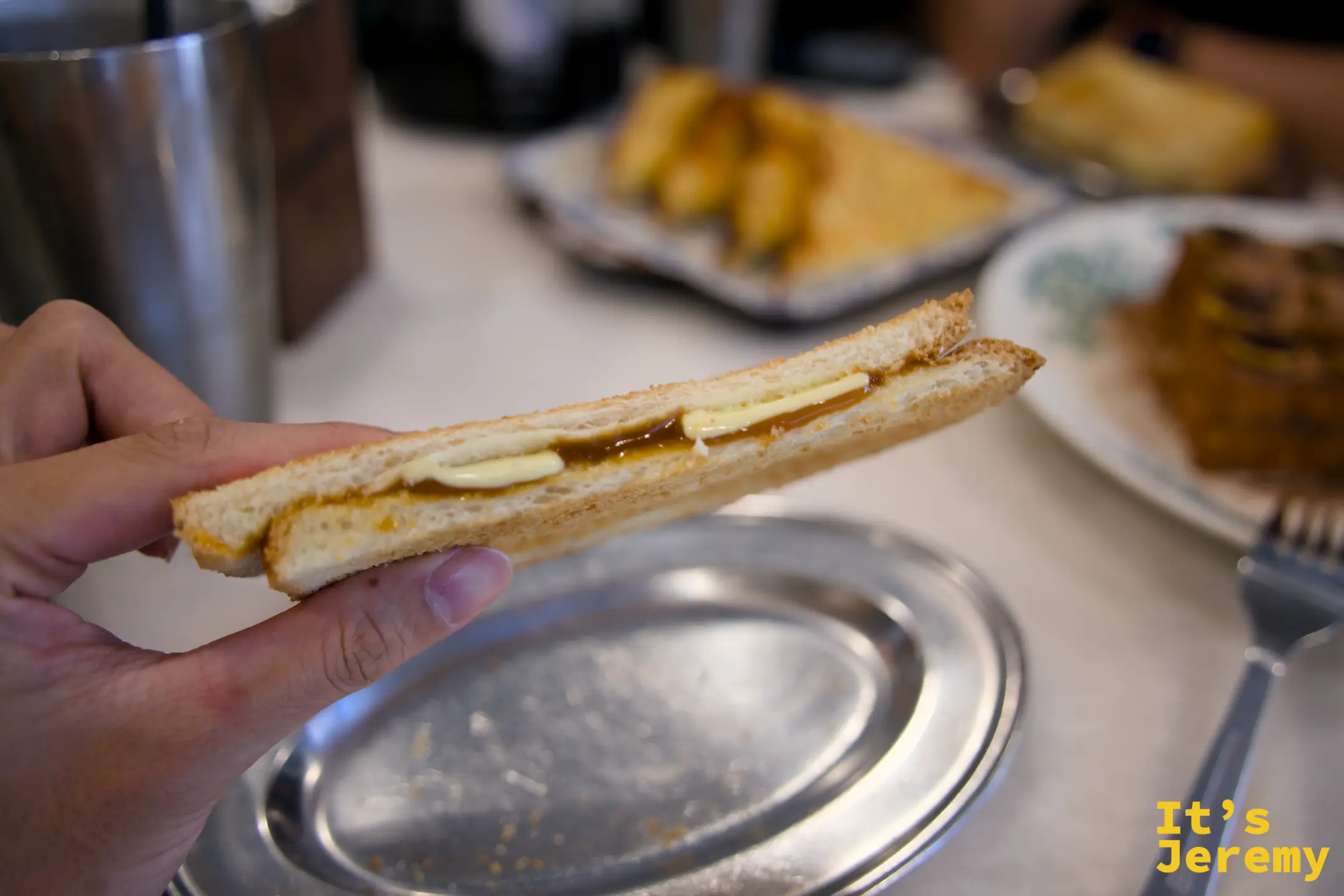 手持剖開的 Kaya Butter Toast，夾層裡的奶油片與椰醬清晰可見，下方是銀色橢圓托盤