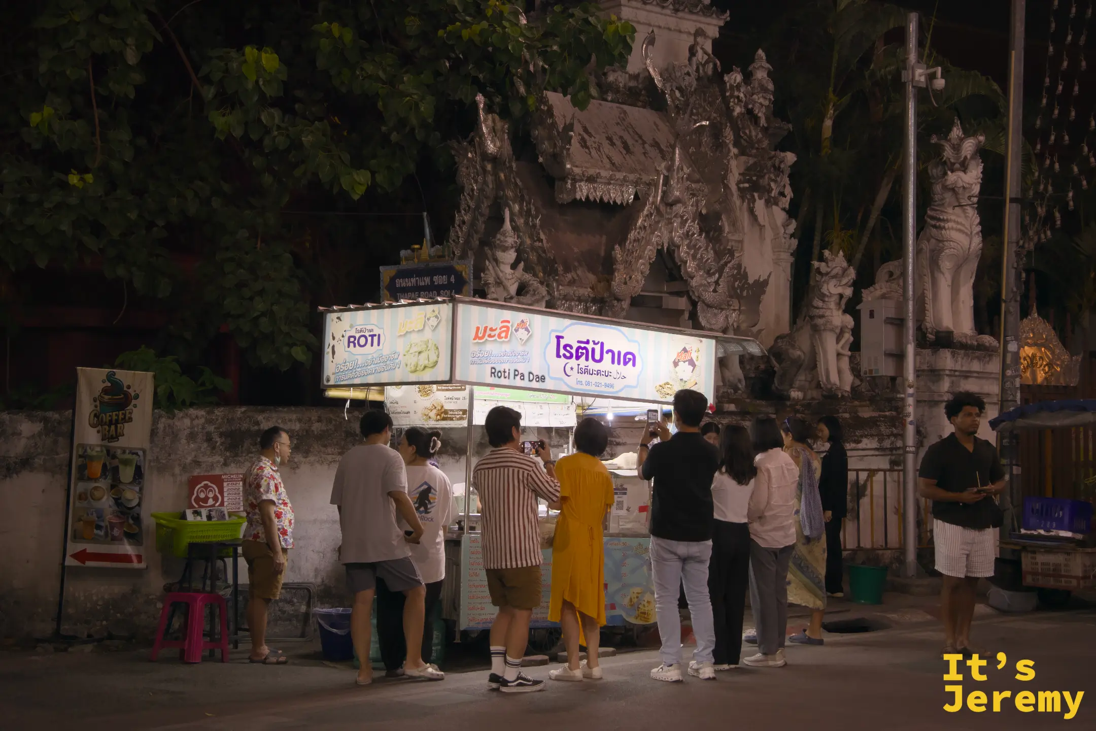 夜裡的 Roti Pa Dae 攤車亮著燈，停在白色寺廟山門前，門前已聚集排隊人潮