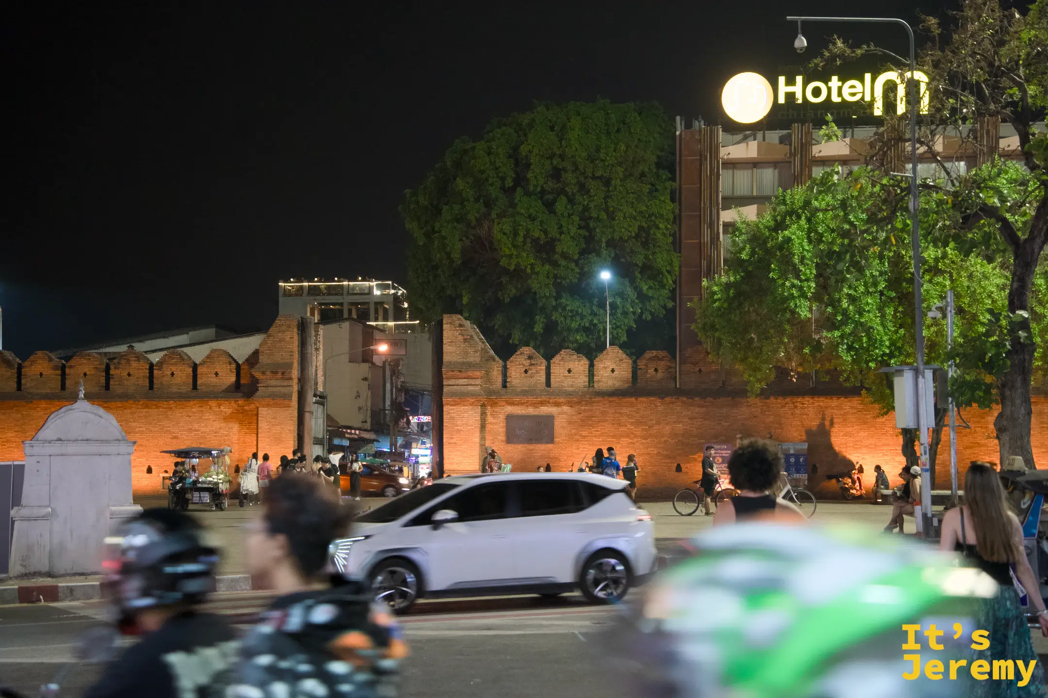 入夜的塔佩門城牆被燈光打成深橘色，城垛輪廓清晰，後方亮著 Hotel M 的招牌，路上車流與行人交錯