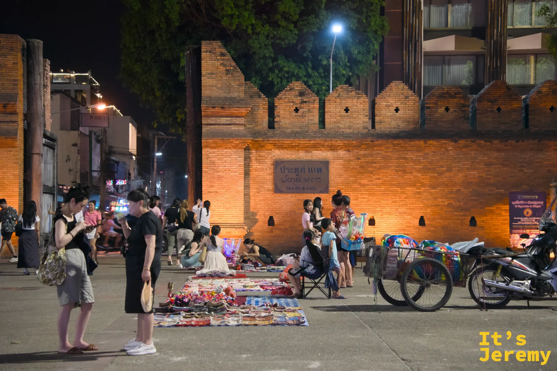 夜裡的塔佩門廣場，攤販在地上鋪布販售織品與小物，遊客在橘色燈光的城牆前走動