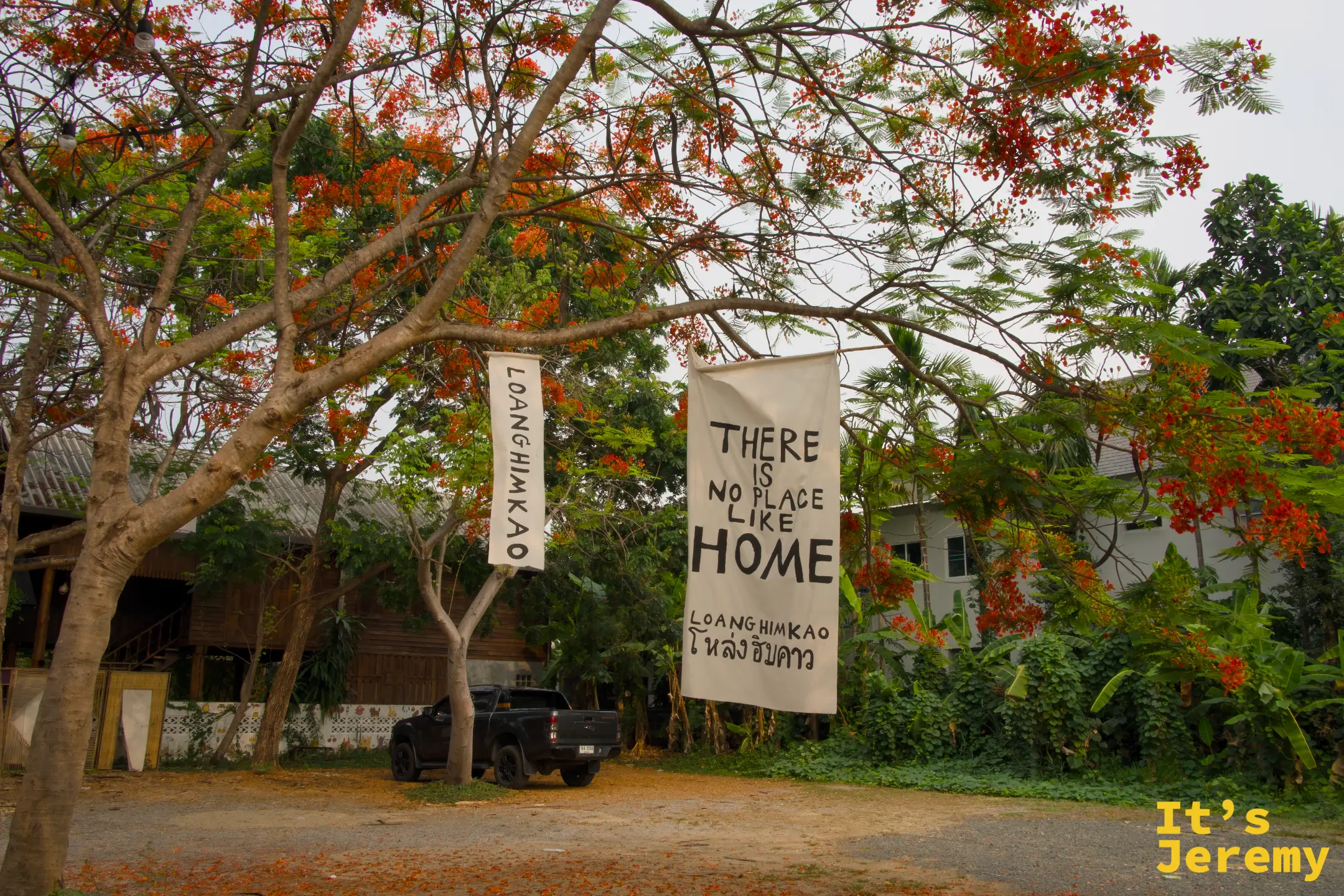 鳳凰花樹下掛著白色大布旗，寫著「THERE IS NO PLACE LIKE HOME — LOANGHIMKAO โหล่งฮิมคาว」