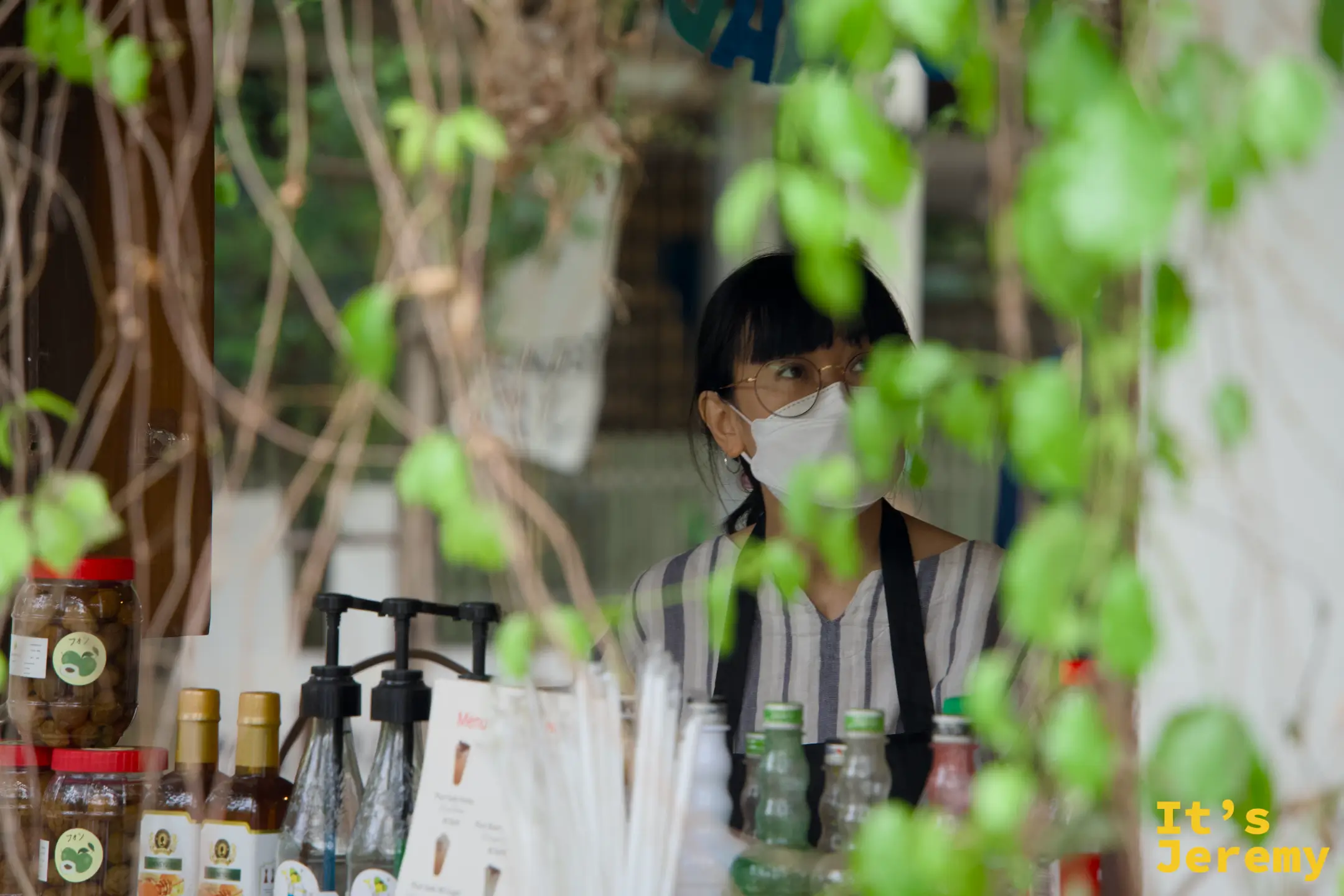 透過藤蔓縫隙看 EDUMade 的老闆娘，戴著眼鏡與口罩站在攤位後方
