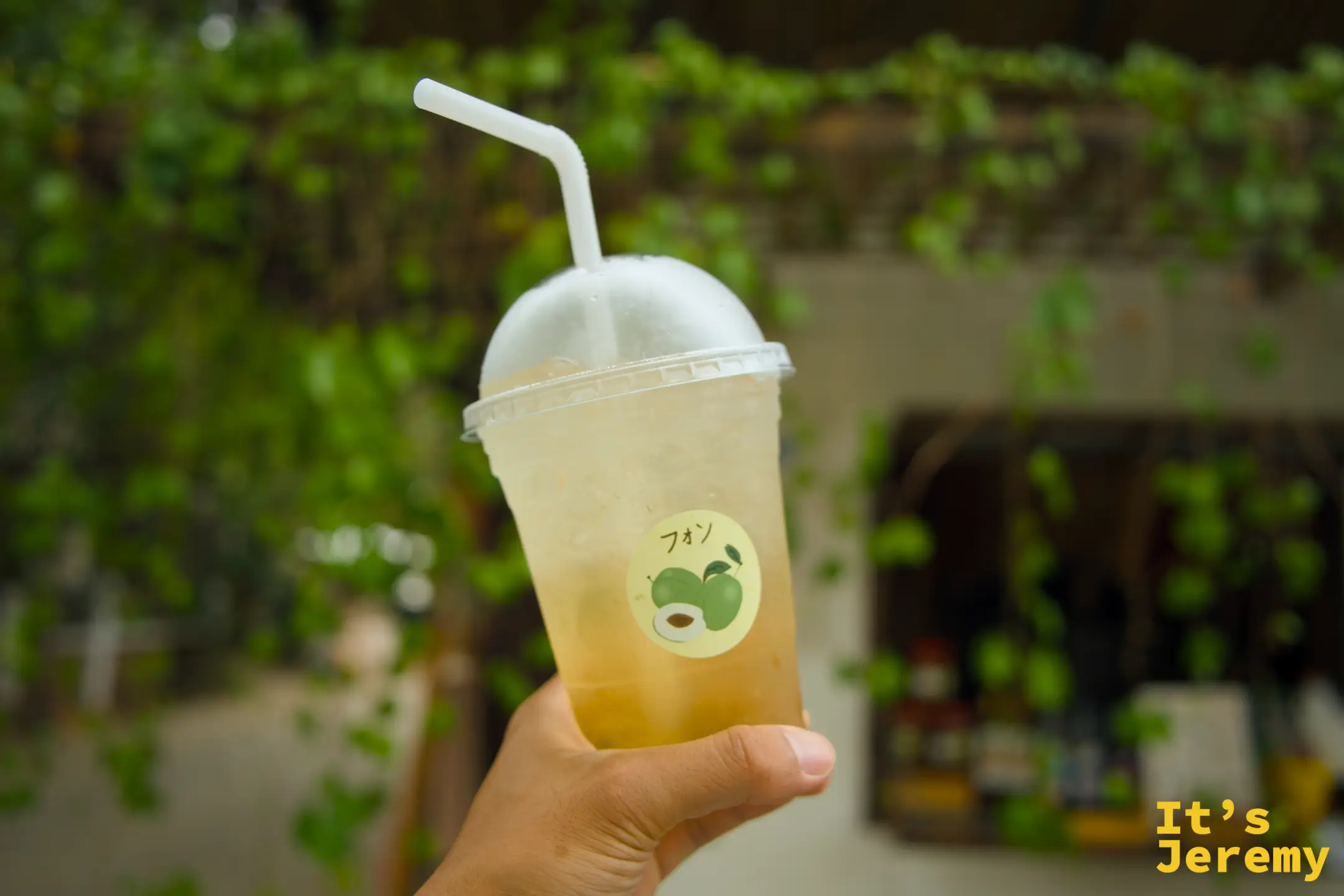 手拿一杯 EDUMade 的梅子蘇打，杯身貼著綠梅貼紙，背景是藤蔓垂落的店面