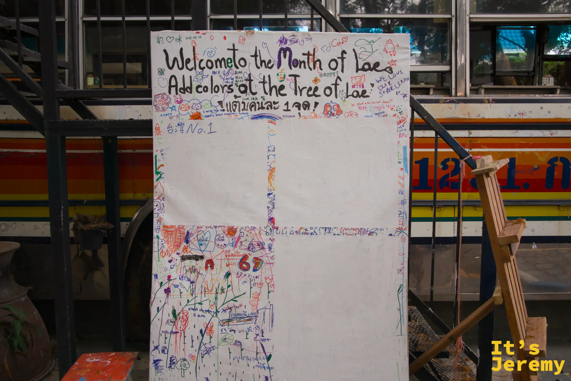 彩色巴士前的白色留言塗鴉板，寫著「Welcome to the Month of Lae, Add colors at the Tree of Lae」，畫滿各色塗鴉